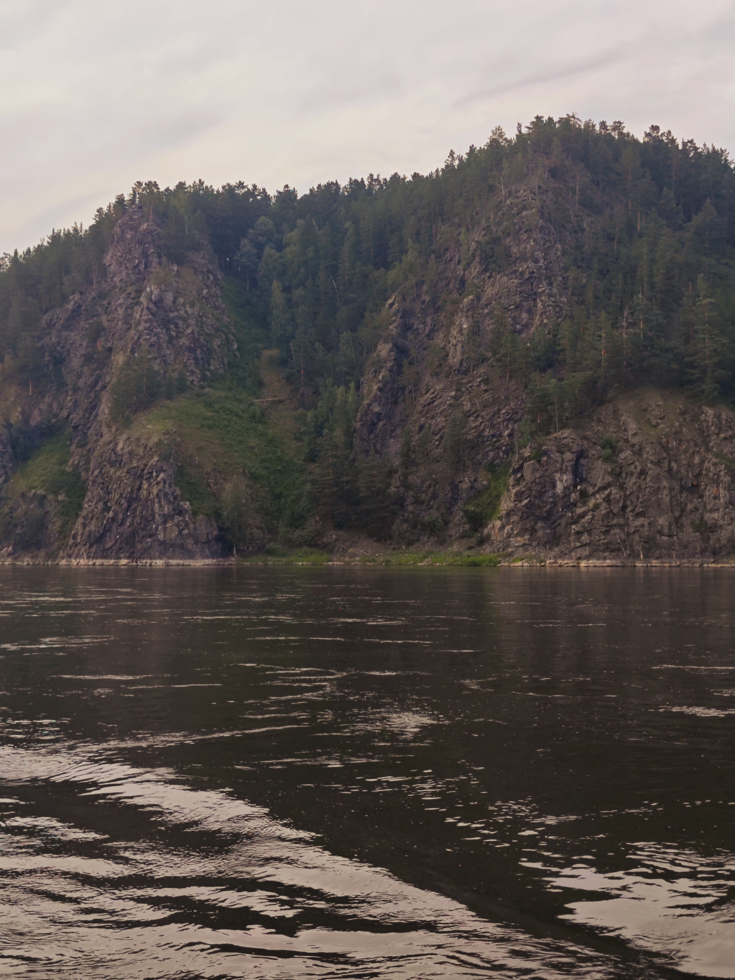 A wide, dark river occupies the foreground of the image, with ripples and waves reflecting the overcast sky. In the middle ground, a steep, rocky cliff face rises directly from the riverbank. The cliff is partially covered in dense green vegetation and scattered trees. The upper portion of the cliff is densely forested with dark green coniferous trees. The sky above is a pale gray, suggesting a cloudy day. The scene appears to be a natural landscape, likely in a rural or wilderness area, with no visible signs of human habitation or activity beyond the implied presence of a river suitable for travel. The location is identified as Beregovaya-Podemnaya, Russia.