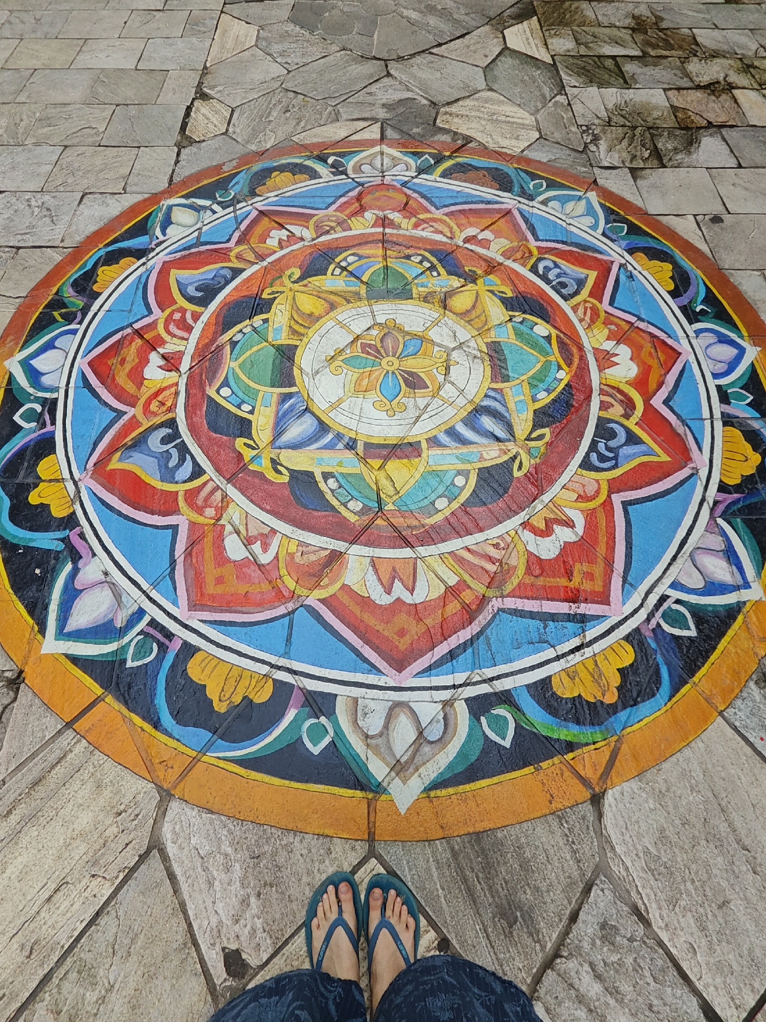 The image shows a colorful, circular mandala painted on a paved ground surface in Pokhara, Nepal. The mandala features intricate geometric patterns and floral motifs in vibrant colors like red, blue, yellow, green, and orange, with black and white outlines. The ground is made of irregular-shaped stone tiles that surround the mandala. At the bottom of the image, a person's feet are visible wearing blue flip-flops and dark blue patterned pants. The scene appears to be outdoors, possibly in a public area like a street or courtyard, given the paving stones and the mandala's presence.