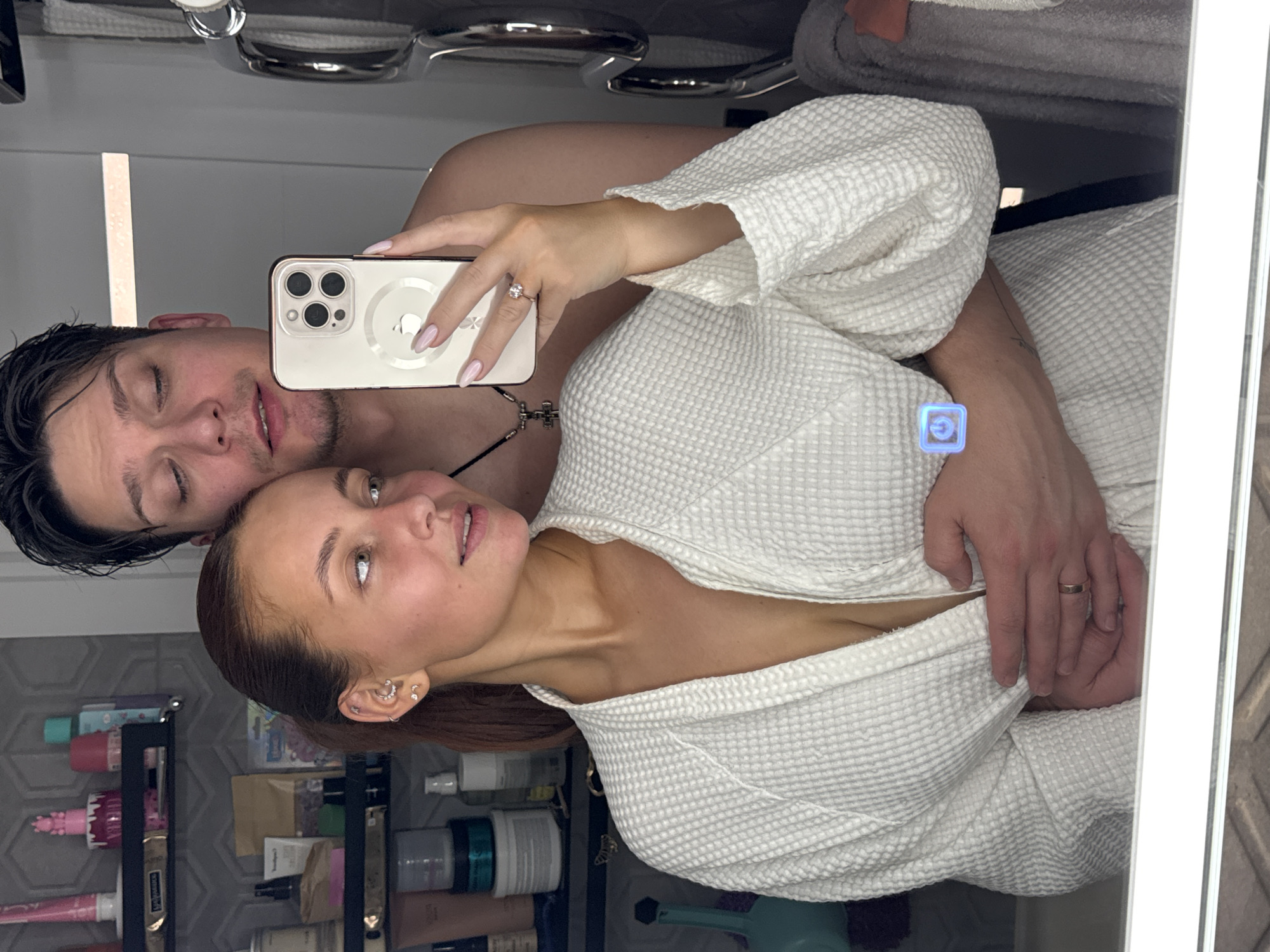 A couple is taking a selfie in a bathroom. The woman is wearing a white bathrobe and has her arm around the man. The man is standing behind her, with his arm around her waist. The woman is holding a phone in her right hand to take the selfie. The woman is wearing a wedding ring on her finger. There are bathroom toiletries and shelves visible in the background, suggesting the location is a bathroom in a house or apartment in Chaikovsky, Russia.