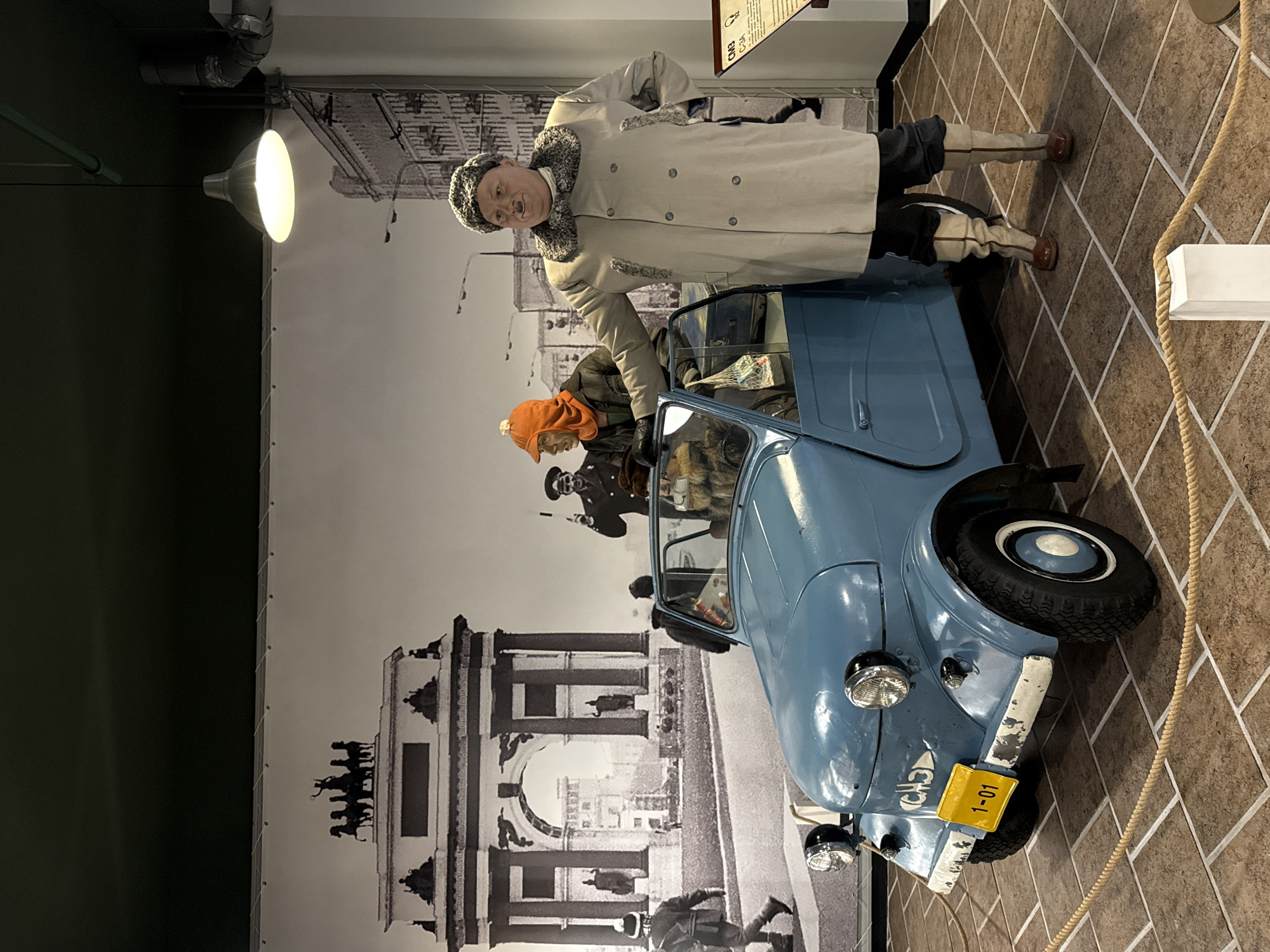 The image depicts a museum diorama set in the city of Arkhangel’skoye, Russia, featuring a vintage light blue car and two mannequins dressed in historical winter attire. The backdrop is a black and white mural of an archway with a horse-drawn carriage atop, and urban architecture.
In the foreground, a light blue, two-door car with its driver's side door open is positioned on a tiled floor. The car has visible headlights, a yellow license plate displaying "1-01", and black tires with white rims.
A mannequin stands to the right of the car, leaning on the open door. This mannequin is wearing a beige, double-breasted coat with fur trim around the collar and down the front. It has dark pants and tall, cream-colored boots with red toe caps. The mannequin has a round face, a flat nose, and a small mustache, and is wearing a fur-lined hat.
Another mannequin is visible inside the car, wearing a green jacket and an orange hood. A bag of groceries is on the seat next to it. A third mannequin, dressed in a dark uniform with a hat, stands behind the car in the mural backdrop, holding a stick.
The environment appears to be indoors, with a high ceiling and a hanging lamp. A rope barrier is present in the foreground, indicating the display is part of an exhibition.