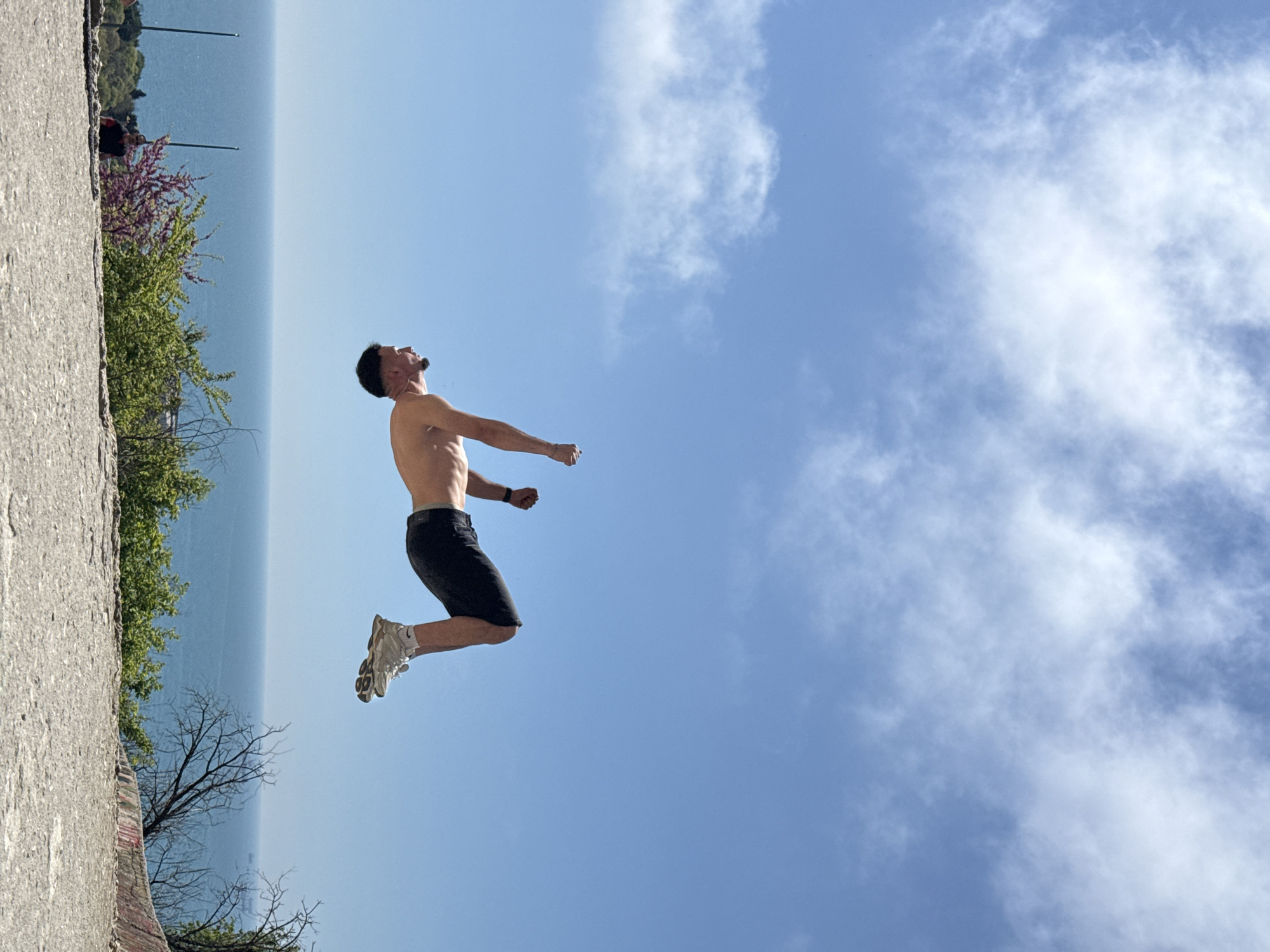 A man is photographed mid-air, leaping near a coastline. He is shirtless, wearing dark shorts and light-colored sneakers. The background features a blue sky with scattered clouds and a sliver of what appears to be the Black Sea. The landscape near the body of water has greenery and a concrete structure. Based on these features, and the distinctive appearance of the coastline, this photograph was taken in Varna, Bulgaria.