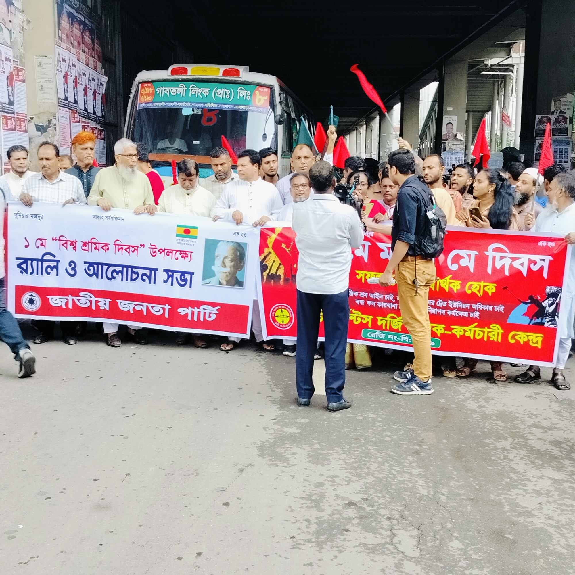 A group of people, predominantly men, are gathered outdoors under what appears to be a concrete overpass, participating in a rally or demonstration. In the background, a white bus from "Gabtoli Link (Pvt.) Ltd." is visible, displaying a route that includes locations like EPZ, Gabtoli, Shyamoli, Farmgate, Shahbag, Motijheel, Sayedabad, Jatrabari, and Signboard, indicating a setting in Bangladesh, likely Dhaka.
The crowd is holding several large banners and flags. Many red flags are visible, some raised high, along with a few green flags. The ground is paved, possibly asphalt, with some wet patches. Posters are plastered on a vertical surface to the left of the image.
The event is identified by the banners as a "Rally and discussion meeting" on the occasion of "1st May 'International Workers' Day'". One white banner with a red border prominently displays "জাতীয় জনতা পার্টি" (Jatiyo Janata Party - National People's Party) and features an image of a political figure. Other text on this banner includes "দুনিয়ার মজদুর" (Workers of the world), "আল্লাহ সর্বশক্তিমান" (Allah the Almighty), and "দেশ কার দেশ কার জনতার জনতার" (Whose country, whose country? The people's, the people's).
A red banner states "মে দিবস সার্থক হোক" (May Day be successful) and lists demands such as "প্রান্তিক শ্রম আইন চাই" (We want a fair labor law), "বন্ধ কল কারখানা" (Closed factories), "ট্রেড ইউনিয়ন অধিকার চাই" (We want trade union rights), and "নিরাপদ কর্মক্ষেত্র চাই" (We want a safe workplace). This banner is associated with the "গার্মেন্টস দর্জি শ্রমিক-কর্মচারী কেন্দ্র" (Garments Tailor Workers-Employees Center).
The participants appear engaged, with some looking serious, and others, with open mouths, suggesting they might be shouting slogans. A person in a white shirt is seen facing away from the camera, seemingly filming or directing, while another individual with a backpack stands observing. The lighting suggests it is daytime.