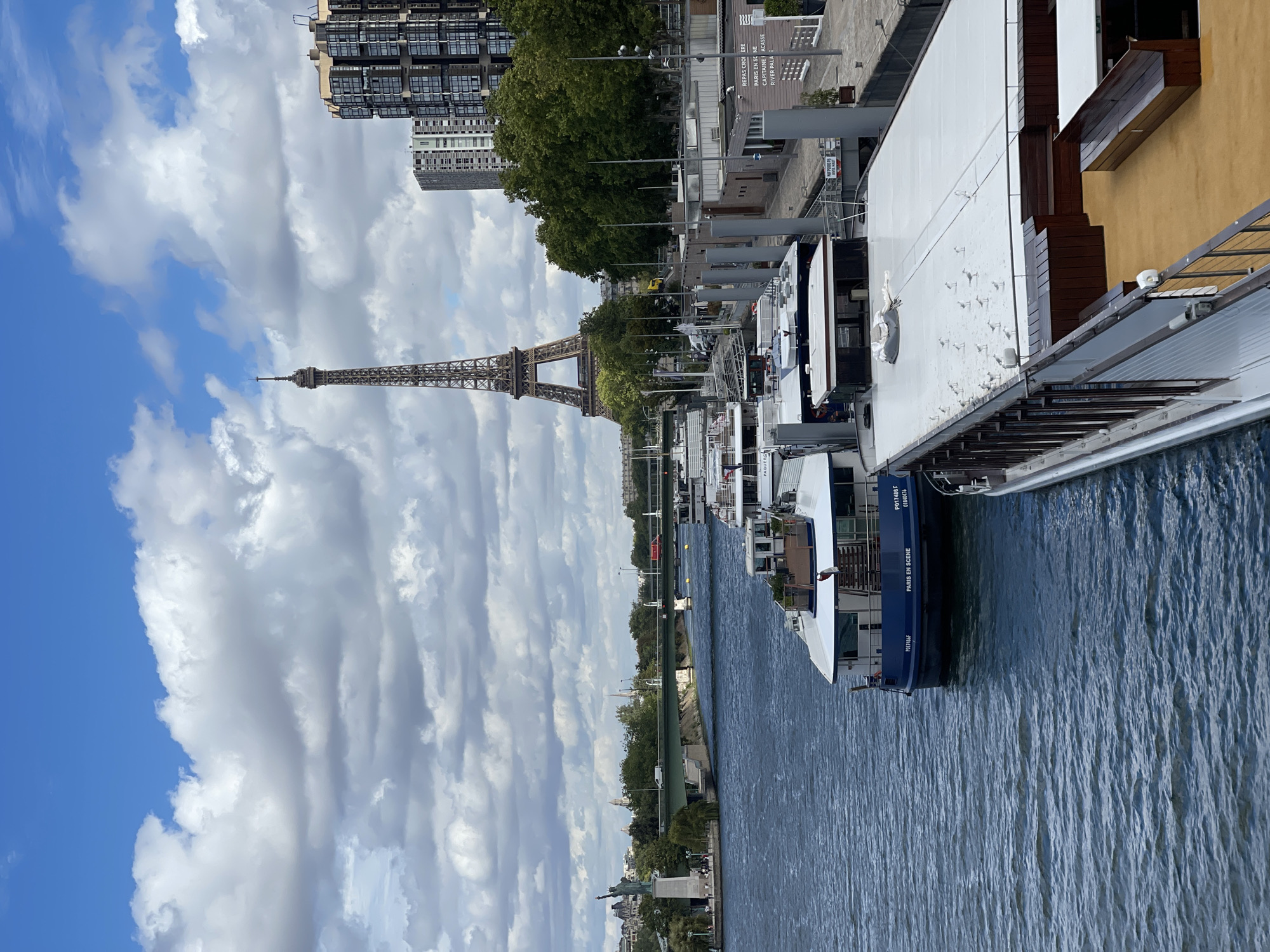 This is a high-angle, medium shot of the Seine River in Paris, France, on a partly cloudy day. In the background, the Eiffel Tower is prominently visible, standing tall against a backdrop of a blue sky with scattered white and gray clouds. To the right of the Eiffel Tower, modern high-rise buildings line the bank, characterized by their dark facades and numerous windows.
In the middle ground, a wide, blue river flows, with several tour boats docked along a walkway on the right side of the frame. The boats vary in size and design, with some appearing to be passenger ferries or cruise vessels. One boat in the foreground, painted dark blue, has "PARIS EN SCENE" printed on its side. The walkway is made of concrete and has a series of grey poles and railings.
Further to the left in the middle ground, a green bridge spans the river. On the left bank, the Statue of Liberty replica on the Grenelle bridge can be seen, with a few pedestrians walking along the riverside promenade. Lush green trees are scattered throughout the scene, softening the urban landscape. The overall impression is a serene urban vista, capturing the iconic landmarks of Paris.