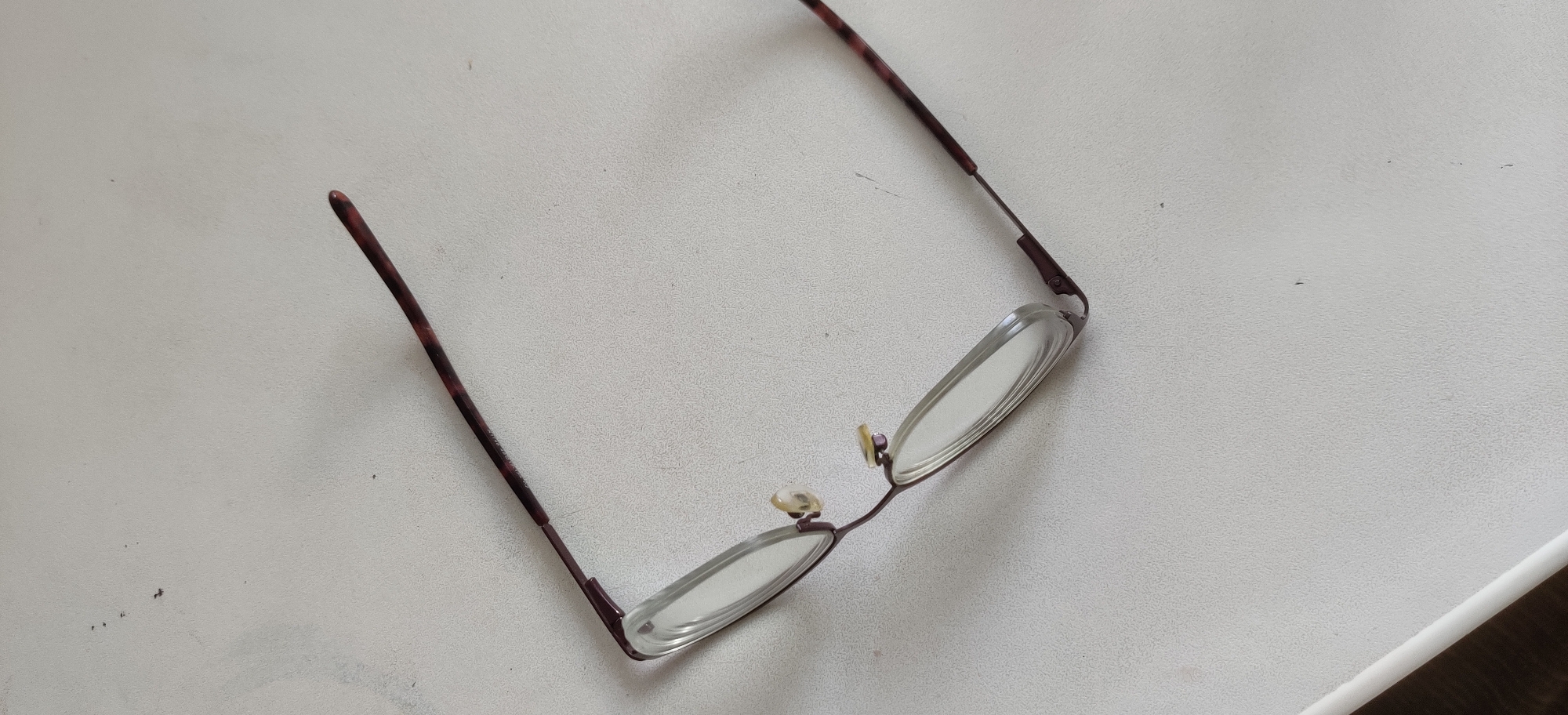 An overhead, eye-level shot captures a pair of eyeglasses lying open on a textured, white surface. The eyeglasses are angled, with the left lens closer to the viewer and the right lens extending away and to the right. The frames are a dark, mottled brown, appearing to be tortoiseshell. The lenses are clear and oval-shaped. The eyeglasses are positioned on a white surface that has a subtle, irregular texture, possibly a table or countertop. There are a few small, dark specks or marks scattered across the surface, and a thin white edge is visible at the bottom of the frame. The lighting is even, casting no significant shadows. The image does not provide any information about the location in Yekaterinburg, Russia.