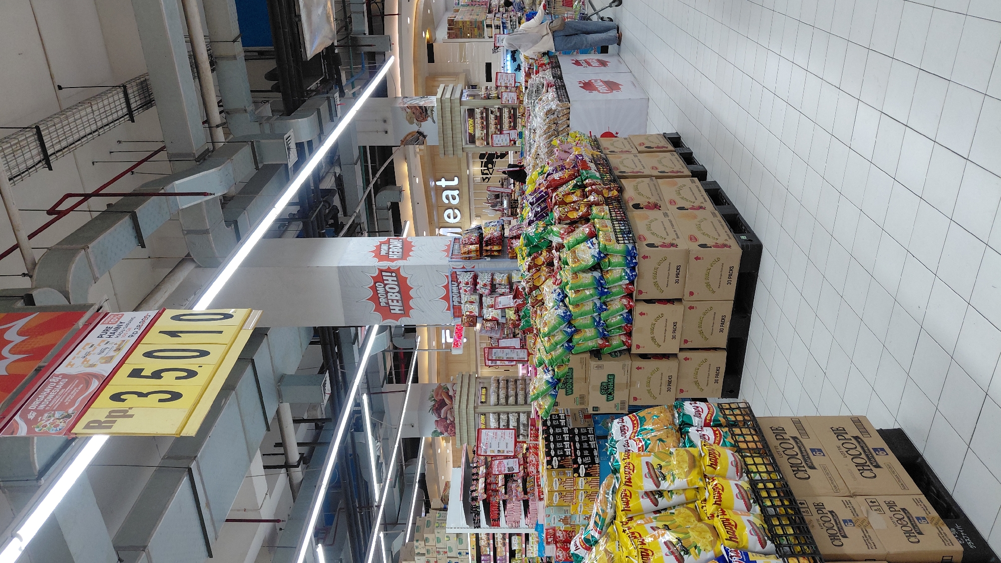 This is a photograph of the interior of a supermarket in Purwokerto, Indonesia. The image is taken from a low angle, looking upwards at a promotional sign hanging from the ceiling. The sign displays a price of "Rp. 35010" in large yellow numerals on a yellow background, with some text above it that is mostly obscured by the image frame. To the right of the price, there is a product label with the words "PURE HANNY MADU JERANG" which translates to "Pure Hanny Orange Honey." Further to the right, the word "Meat" is visible in large white letters on a dark background, indicating a meat section.
In the foreground, there are several aisles of merchandise, primarily stacked boxes of snack products. One notable stack of boxes features the brand "LOTTE CHOCO PIE" in prominent lettering. To the right of these boxes, a display of bagged snacks is visible, with many bags featuring green and yellow packaging. A woman wearing a light-colored hijab and blue pants is seen pushing a shopping cart in the mid-ground, on the right side of the image. The supermarket floor is tiled with white squares. The ceiling features extensive industrial ductwork and lighting. The overall impression is a well-stocked retail environment with promotional displays.