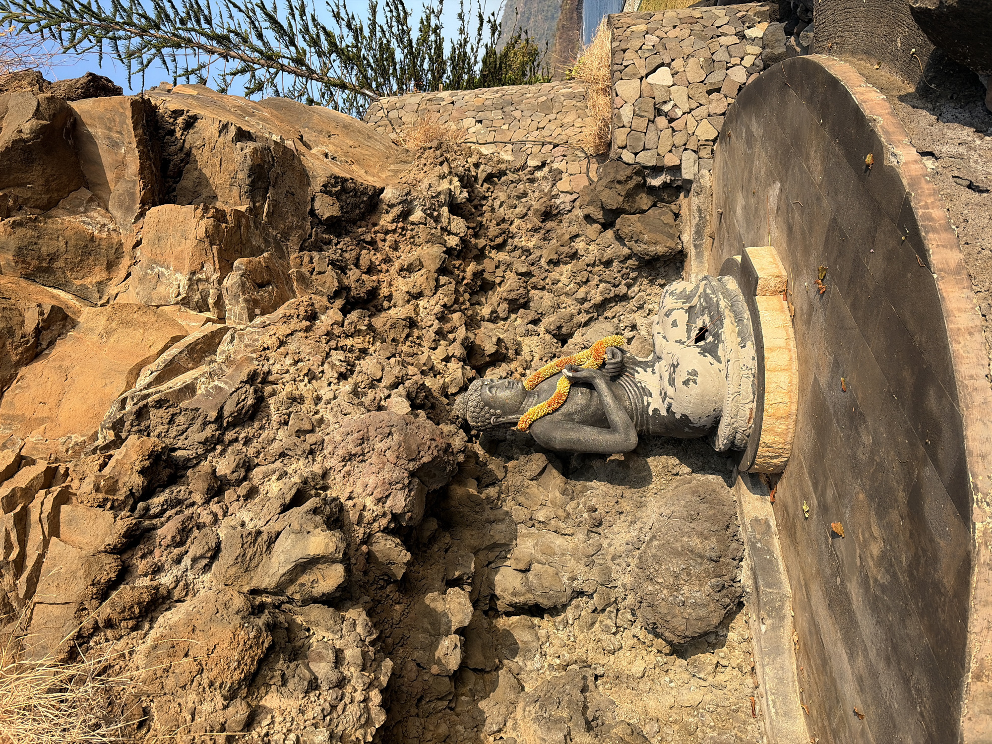 An outdoor stone statue of Buddha is seated at the base of a large rock formation. The Buddha statue is wearing a garland of yellow and orange flowers around its neck. The rock formation is rough and textured, with some areas appearing eroded. To the right of the statue, there is a stone wall and a tall, thin evergreen tree. In the background, a body of water is visible under a clear blue sky. The scene is lit by sunlight, casting shadows on the ground. There is no indication of the specific location within the city of Abang, Indonesia.