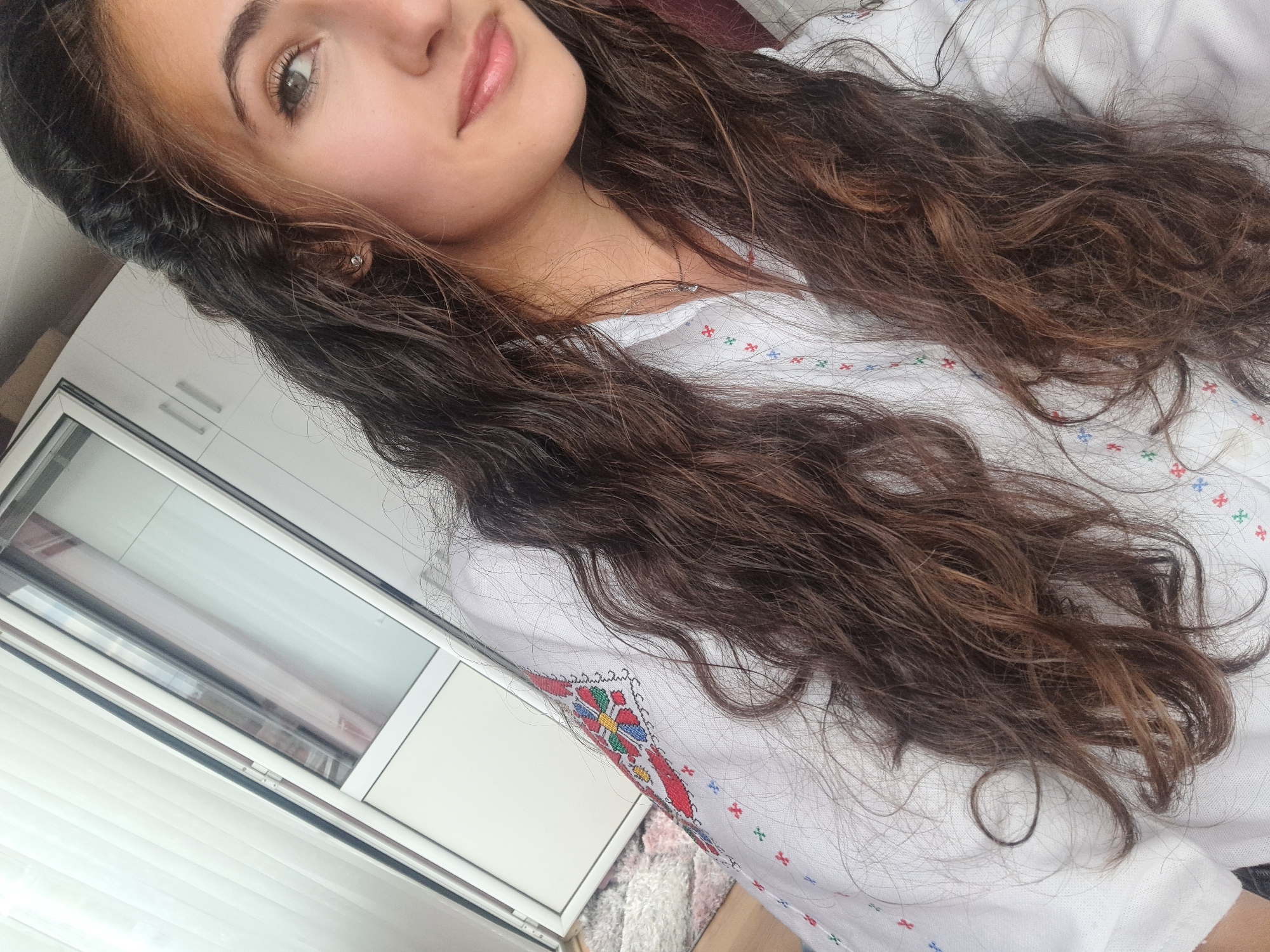 A selfie shot shows a young woman with long, wavy brown hair partially obscuring a white shirt decorated with red, blue, green, and yellow cross-stitch embroidery. She is close to the camera, with part of her face visible, including her eyes, nose, and mouth. A small earring is visible on her left ear. Behind her is a white window frame. The location is Unknown, Unknown.