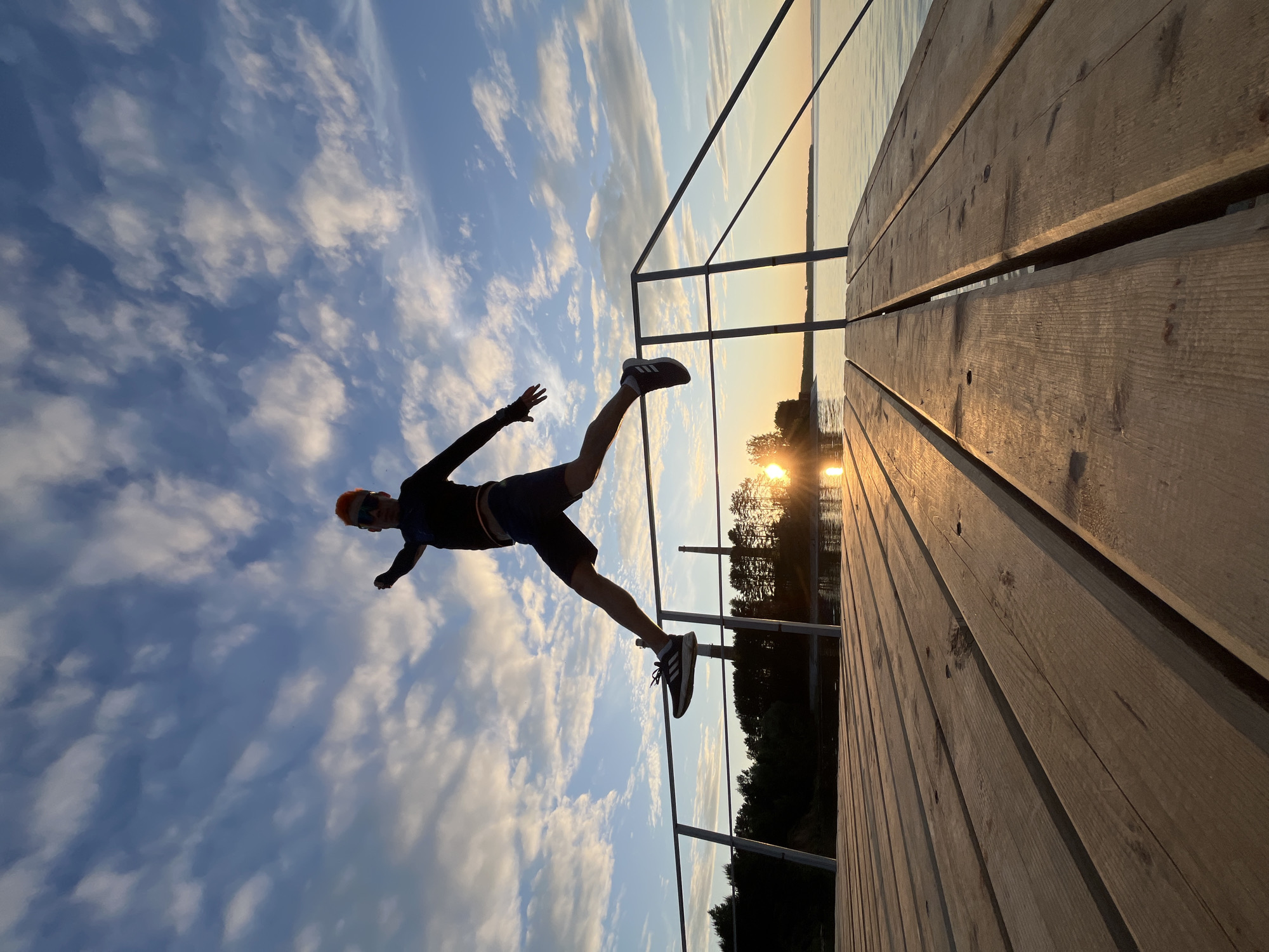 A person is captured mid-jump above a wooden pier, silhouetted against a vibrant sunset sky over a body of water in Chaikovsky, Russia. The individual wears a dark long-sleeved top, shorts, and athletic shoes with white stripes. Their head features bright orange hair or a cap, and they are wearing sunglasses. Both arms are extended horizontally, and legs are spread, one forward and one backward, indicating a dynamic leap. The wooden pier, composed of light-colored planks, extends into the lower-right foreground. A metal railing runs along the right side of the pier. The setting sun, a bright orange disc, is visible low on the horizon towards the right, casting a golden reflection on the calm water and illuminating the undersides of scattered clouds in the blue sky. A dark tree line is visible along the left shoreline, and a tall, slender industrial chimney stands in the mid-ground behind the trees and railing on the left.