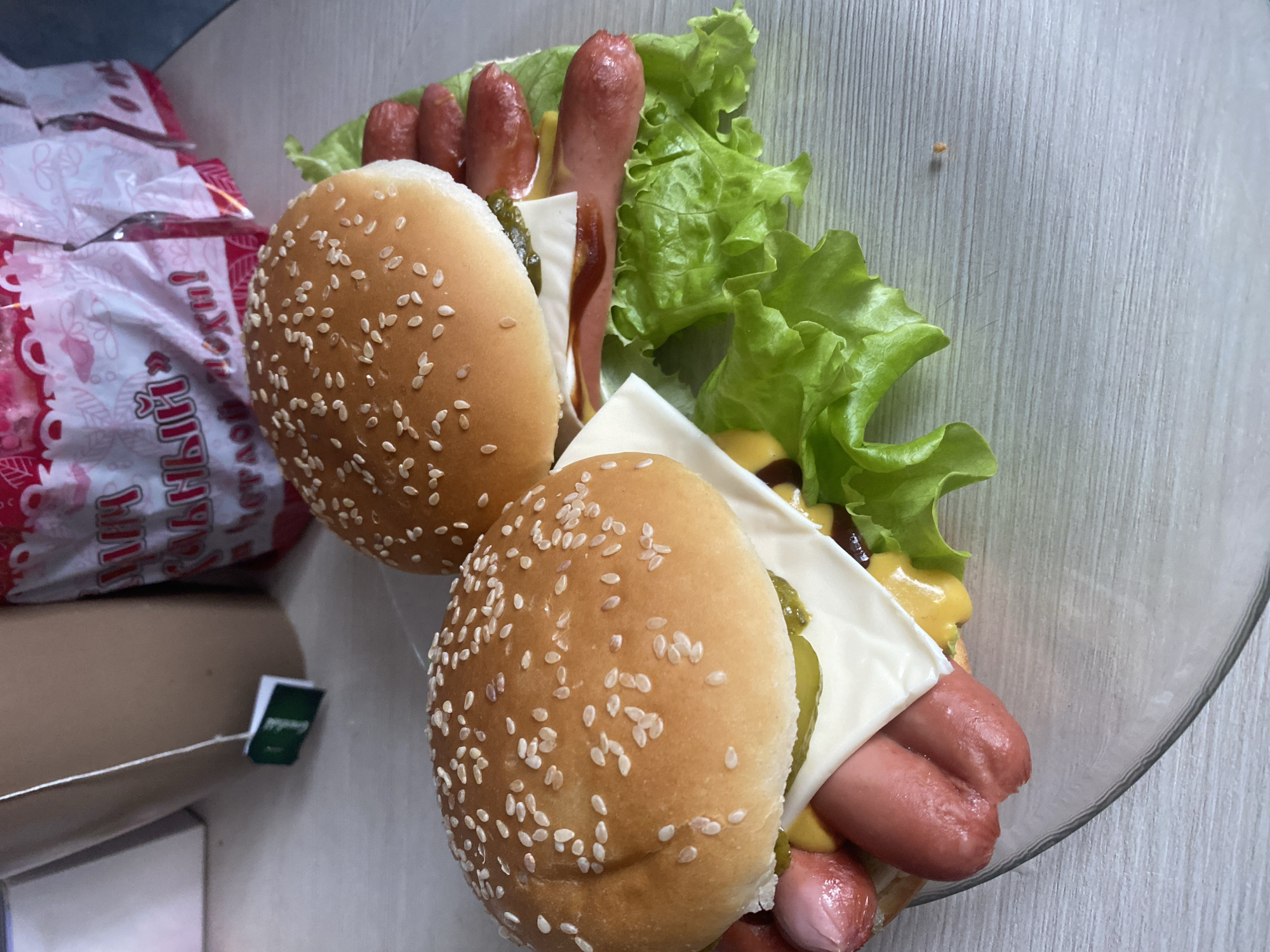Two hot dogs in sesame seed buns, containing lettuce, cheese slices, pickles, mustard, ketchup, and sausages, are positioned in a clear glass bowl. The bowl rests on a gray-toned surface resembling a wooden table. Adjacent to the bowl is a packaged item with red and white labeling, alongside a tea bag with a visible green tag. The location is potentially Izhevsk, Russia, given the presence of Russian labeling on the packaged item.
