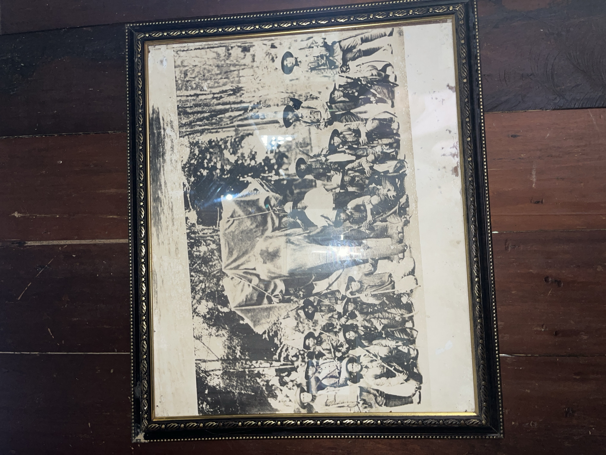 The image is a black and white photograph framed with a decorative black and gold border, displayed against a background of dark wooden planks. The photograph itself depicts a group of approximately 20 individuals, primarily men, posing with what appears to be an elephant covered by a large tarp. The people are dressed in what seem to be military or safari attire, including hats and long-sleeved shirts and pants. Many of the men are holding rifles or spears. They are arranged in rows, with some standing and others kneeling. The elephant is a prominent subject, positioned behind the group. The background suggests an outdoor environment, likely a jungle or forested area, with trees visible. The overall scene conveys an expeditionary or colonial context.