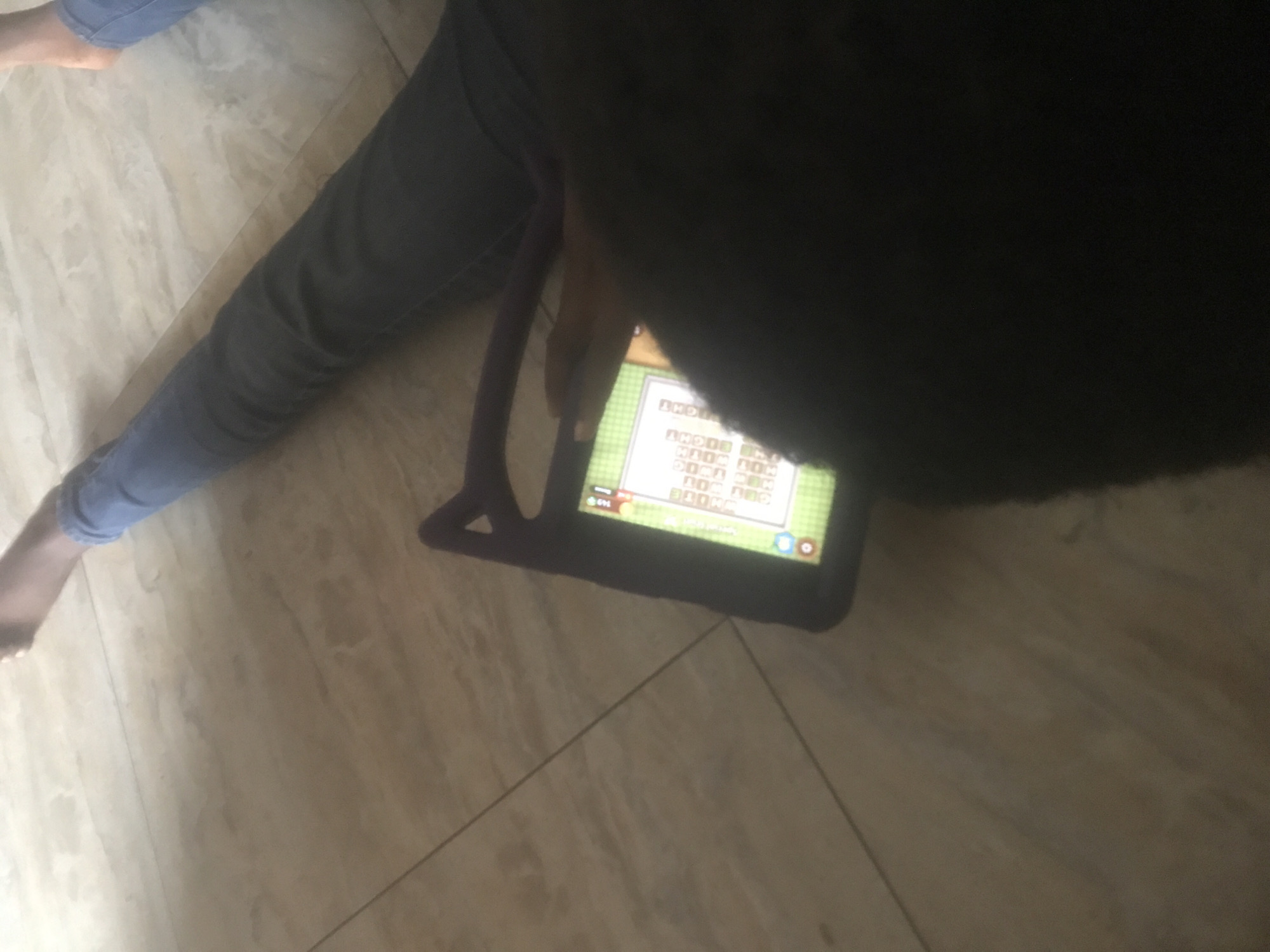The scene depicts at least one individual, partially obscured by shadow, sitting or reclining on a light-colored tiled floor. A person, whose upper body is out of frame or heavily shadowed, wears dark grey or black fitted pants on their left leg and holds a tablet device with their right hand. Their bare right foot is partially visible at the upper right. A separate bare foot and a portion of a leg in light blue jeans are also visible at the far upper right, indicating a second person or a relaxed posture of the first. The tablet, encased in a dark purple protective cover featuring prominent ear-like protrusions, displays a pixelated video game with a light green background, white text boxes, numerical values (e.g., "100," "200"), and various game icons. The floor consists of light beige or light brown rectangular tiles with subtle veining, characteristic of natural stone, and a dark grout line runs diagonally across the mid-left of the frame. The area is illuminated by ambient light, casting shadows in the foreground.