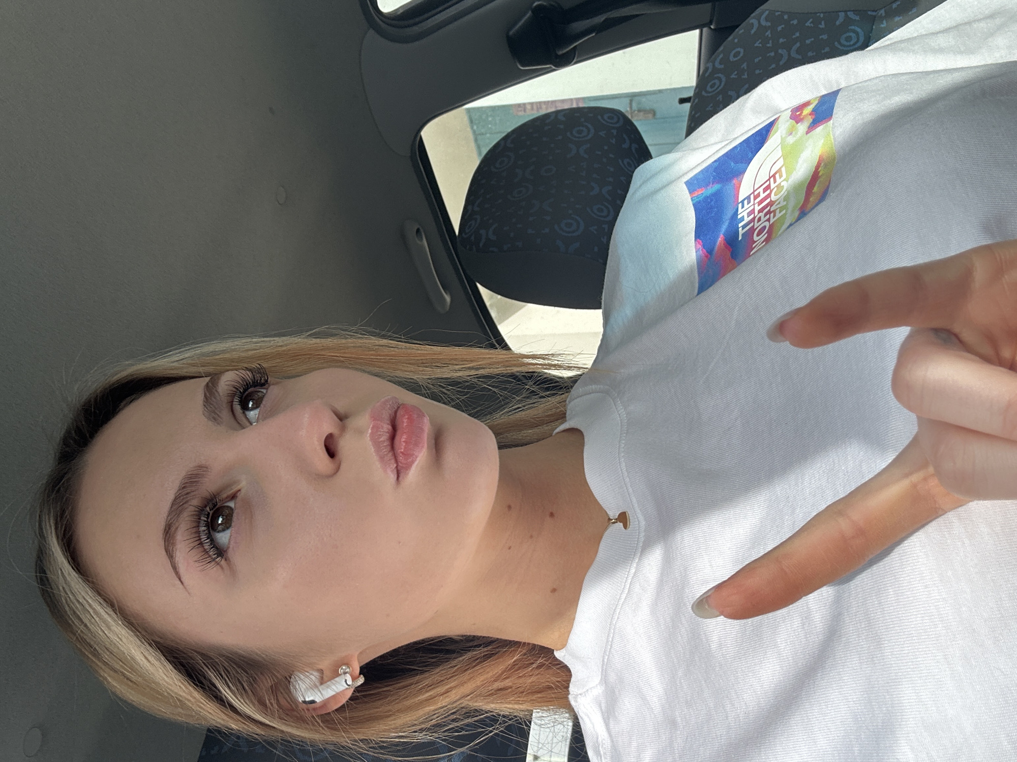 A young woman is seated in a car. She has long blonde hair, false eyelashes, and is wearing wireless headphones. She is wearing a white t-shirt with a colorful "The North Face" logo on the left side of her chest. She is holding up her index and middle finger with her hand. The car interior is gray, and the headrest of the seat behind her is patterned. The location is Unknown, Unknown.
