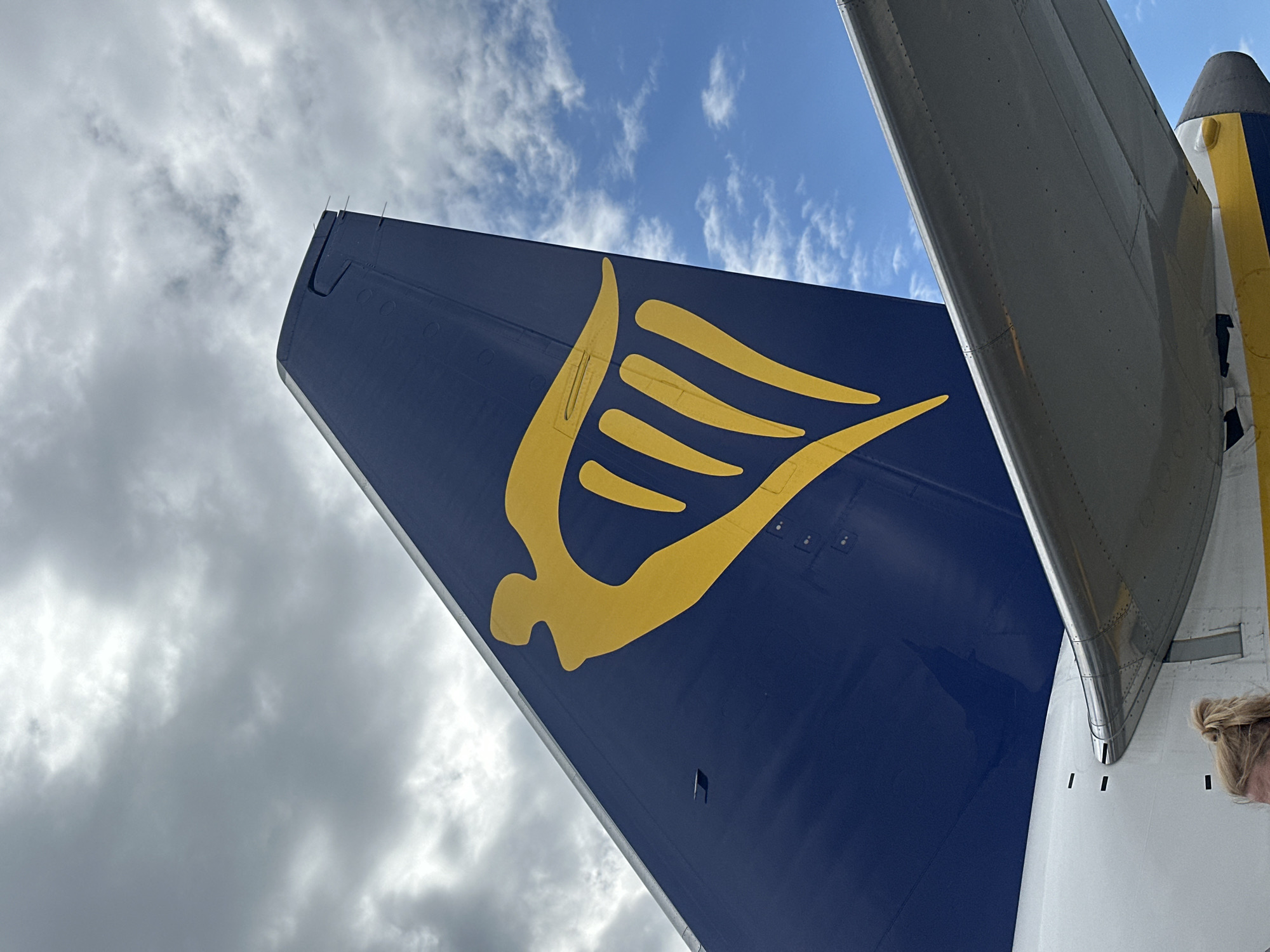 The image captures a close-up view of the tail fin of a Ryanair airplane, prominently displaying the airline's yellow harp logo against a dark blue background. The tail fin is positioned against a sky filled with a mix of white, fluffy clouds and darker, overcast clouds. Below the tail fin, a portion of the airplane's wing is visible, appearing metallic gray and reflecting some of the sky's light. A section of the airplane's fuselage, painted white with yellow and blue stripes, is also visible at the bottom of the frame. A woman with blonde hair tied up in a bun is partially visible at the very bottom of the image, suggesting the photo was taken from ground level. The scene is consistent with an airport environment, and given the detail, it is likely taken at John Paul II International Airport Kraków–Balice (KRK) in Balice, Poland.