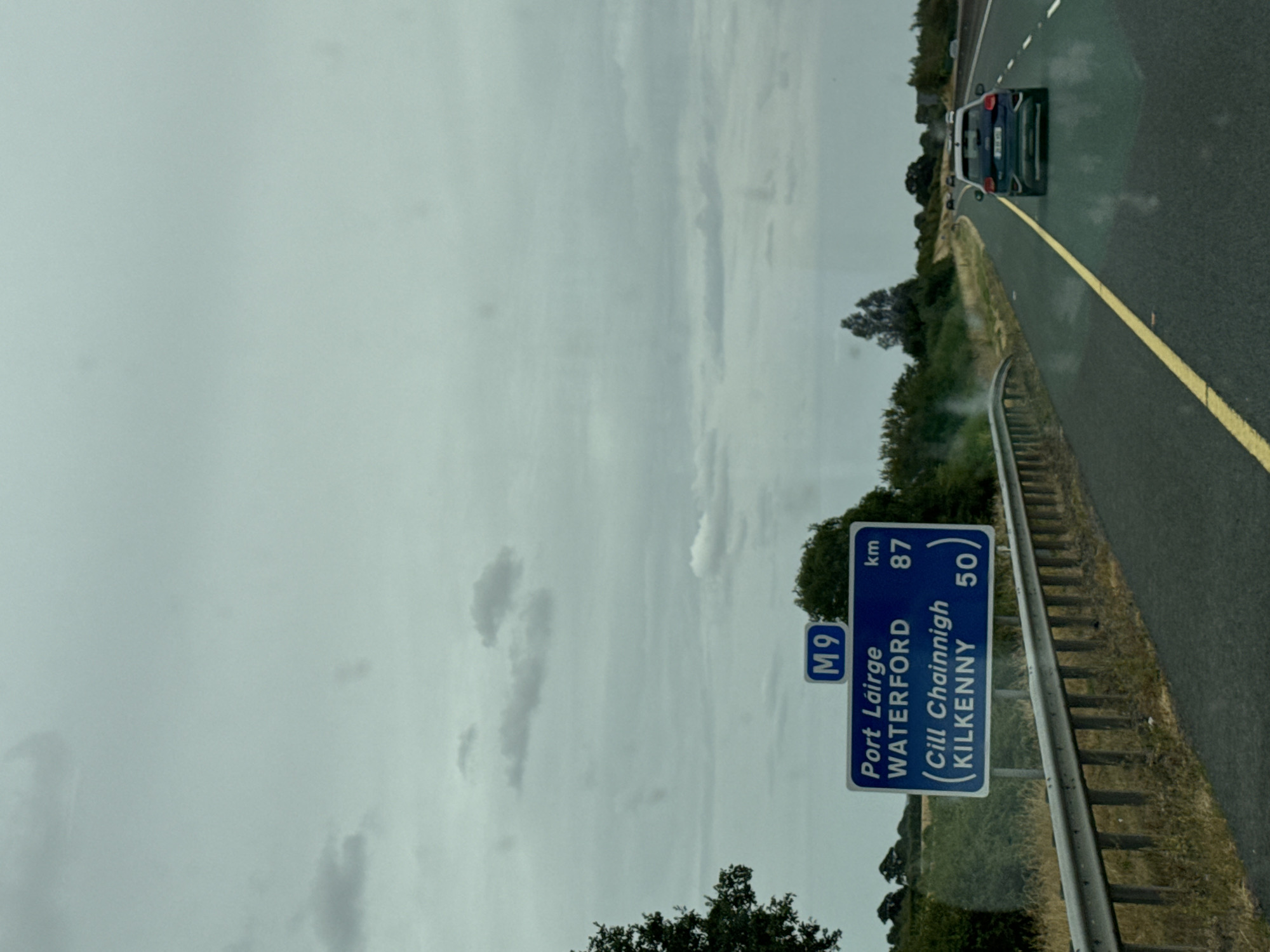 An eye-level, medium outdoor shot captures a highway scene with a blue and white road sign indicating directions and distances. The sign shows "M9" at the top, followed by "Port Lairge WATERFORD" with "km 87" to the right, and in parentheses "(Cill Chainnigh KILKENNY)" with "50" to the right. The sign is positioned next to a metal guardrail. Beyond the guardrail, a grassy verge leads to more vegetation and trees. The road ahead is paved, with a yellow line marking the lane. A dark-colored car is visible driving on the highway. The sky is overcast with grey clouds. The location is identified as Ballyburn, Ireland.