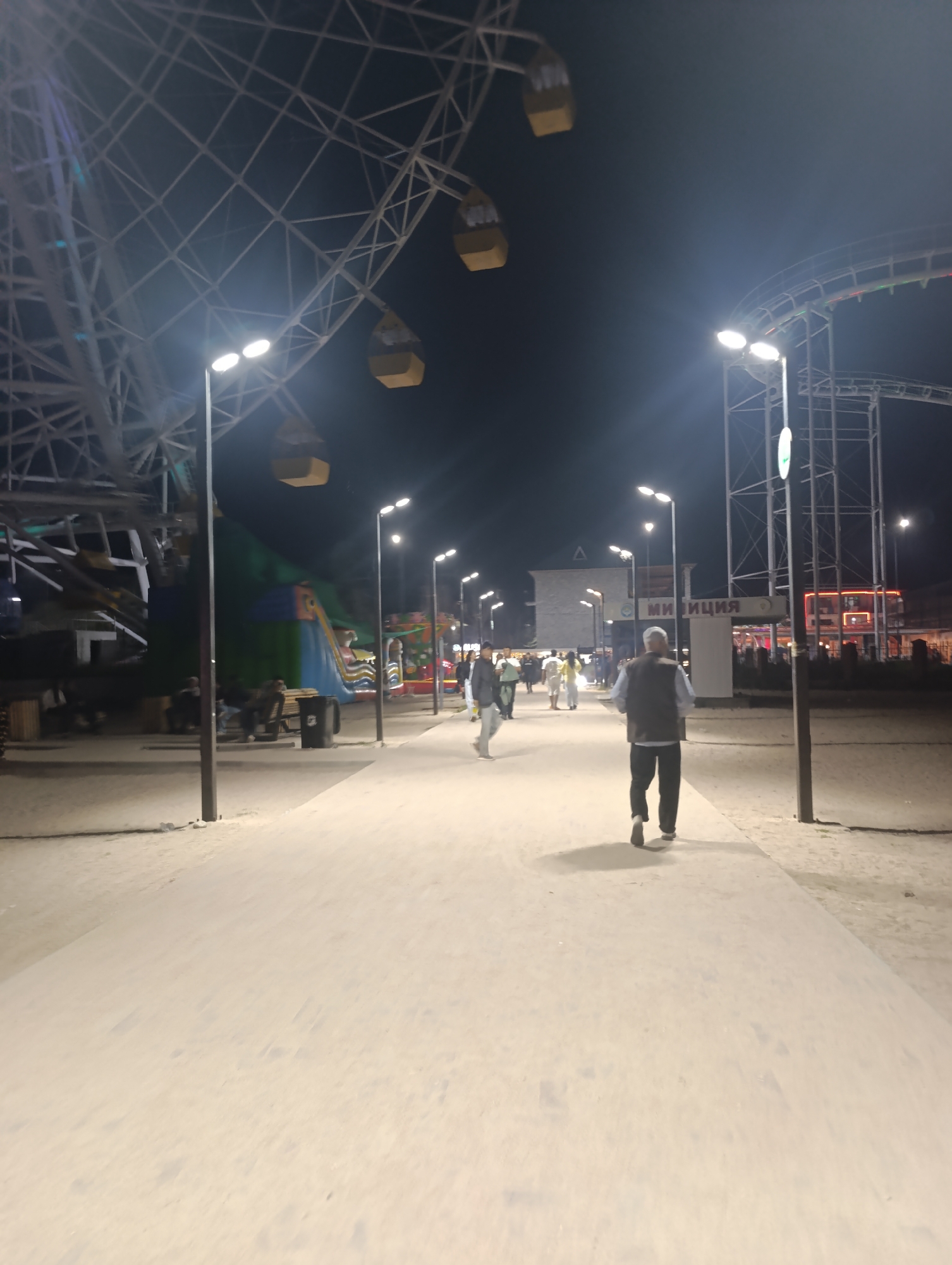 The image depicts a nighttime scene at what appears to be an amusement park or fairground in Bosteri, Kyrgyzstan. A large Ferris wheel dominates the left side of the frame, its metal structure illuminated by ambient light. To the right of the Ferris wheel, a roller coaster track is visible against the dark sky. The ground is a light-colored, sandy or dusty surface, with a paved walkway in the foreground. Several lampposts line the walkway, casting pools of light.
In the mid-ground, a group of people are walking and standing. One individual in the foreground, an older man wearing a dark vest over a light shirt and dark pants, is walking away from the camera. Other individuals, including families and couples, are scattered throughout the scene, some engaged in conversation or movement.
Behind the walkway, various amusement park attractions are visible. A large, inflatable structure, possibly a bouncy castle or slide, is present. Buildings with illuminated windows suggest shops or food stalls. A sign with Cyrillic lettering, reading "МИЛИЦИЯ," is prominently displayed on a structure in the mid-ground, indicating a police or security presence. The overall atmosphere is that of a lively evening at an outdoor entertainment venue.