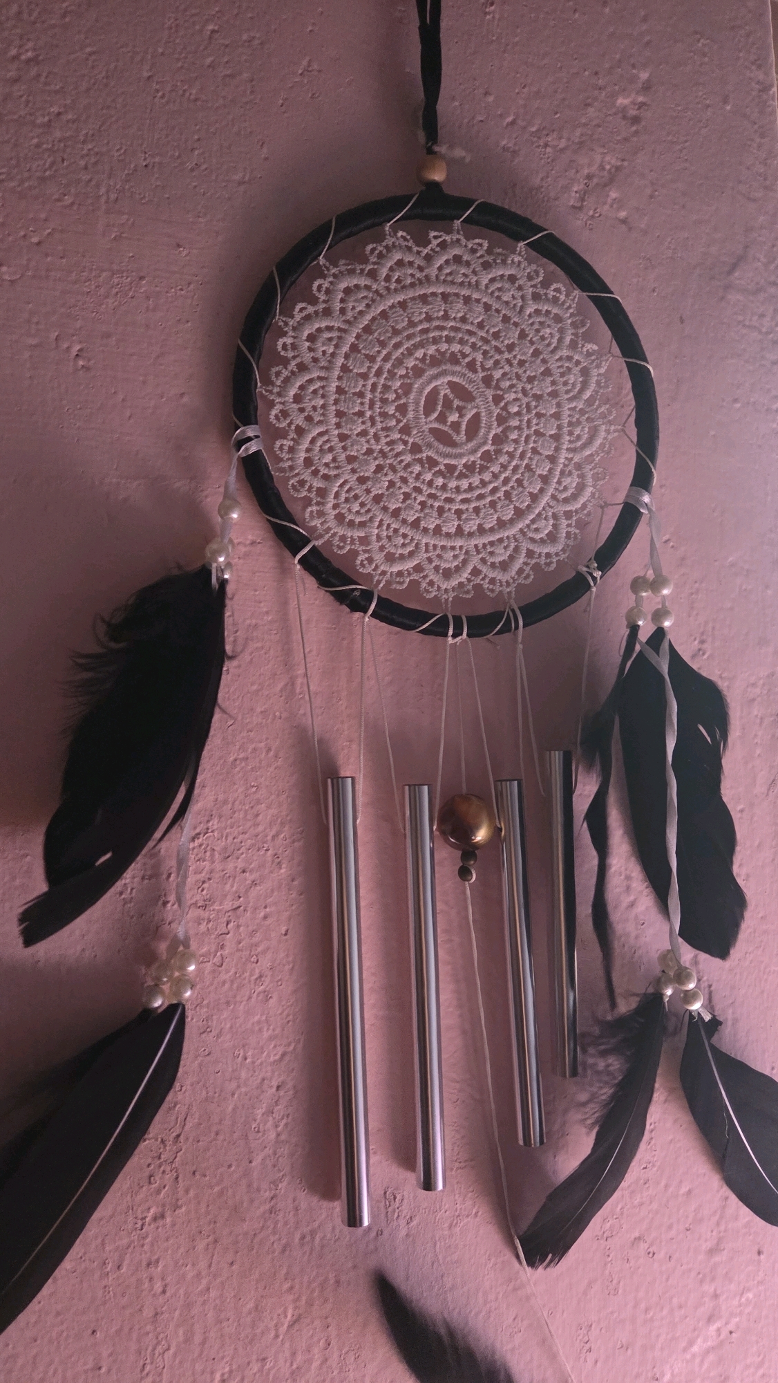 A dreamcatcher with black feathers and silver chimes hangs on a pink, textured wall. The dreamcatcher has a black hoop and a white, lacy center. There are multiple strands hanging from the hoop, some adorned with white beads and black feathers, and others with the silver chimes. One chime is slightly larger than the others and has a metallic, golden-brown color. The background wall appears to be an interior surface. The image is taken from a slightly upward angle, focusing on the dreamcatcher.