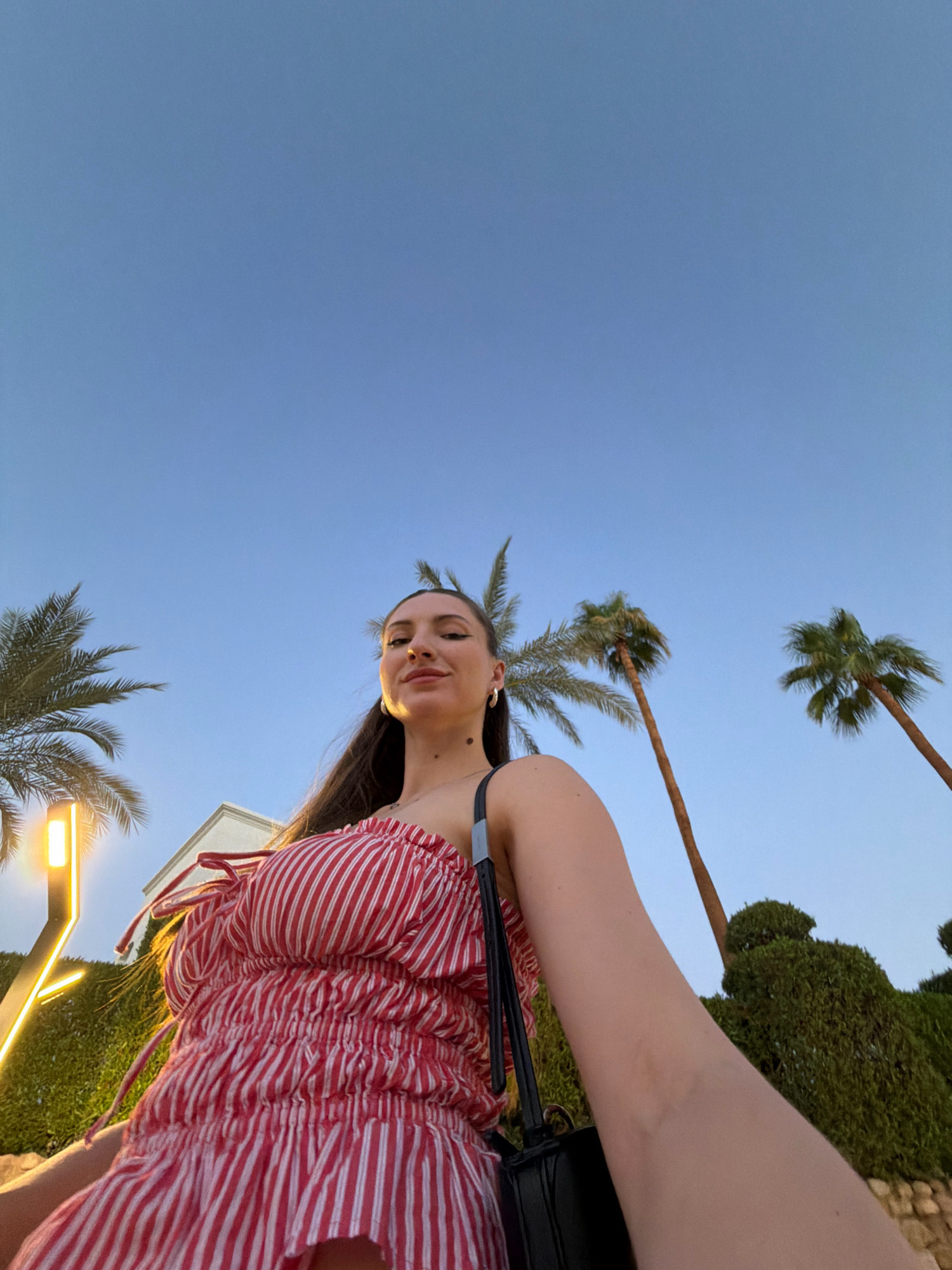 An outdoor, low-angle, eye-level selfie is captured under a clear blue sky, suggesting a warm climate. The subject, a woman, is in the foreground, her face tilted slightly upwards, and she is smiling. She has long, dark hair that falls past her shoulders. She is wearing a red and white striped, off-the-shoulder top with ruching detail. A black purse strap is visible over her right shoulder.
The background features lush green hedges and the silhouettes of palm trees, consistent with a tropical or subtropical location. To the left, a modern outdoor lamp with a yellow light source is visible. Behind the woman, a building with white walls and a peaked roof can be seen. The presence of palm trees and the overall aesthetic strongly suggest a location in Sharm El Sheikh, Egypt, a resort city known for its beaches and warm weather.