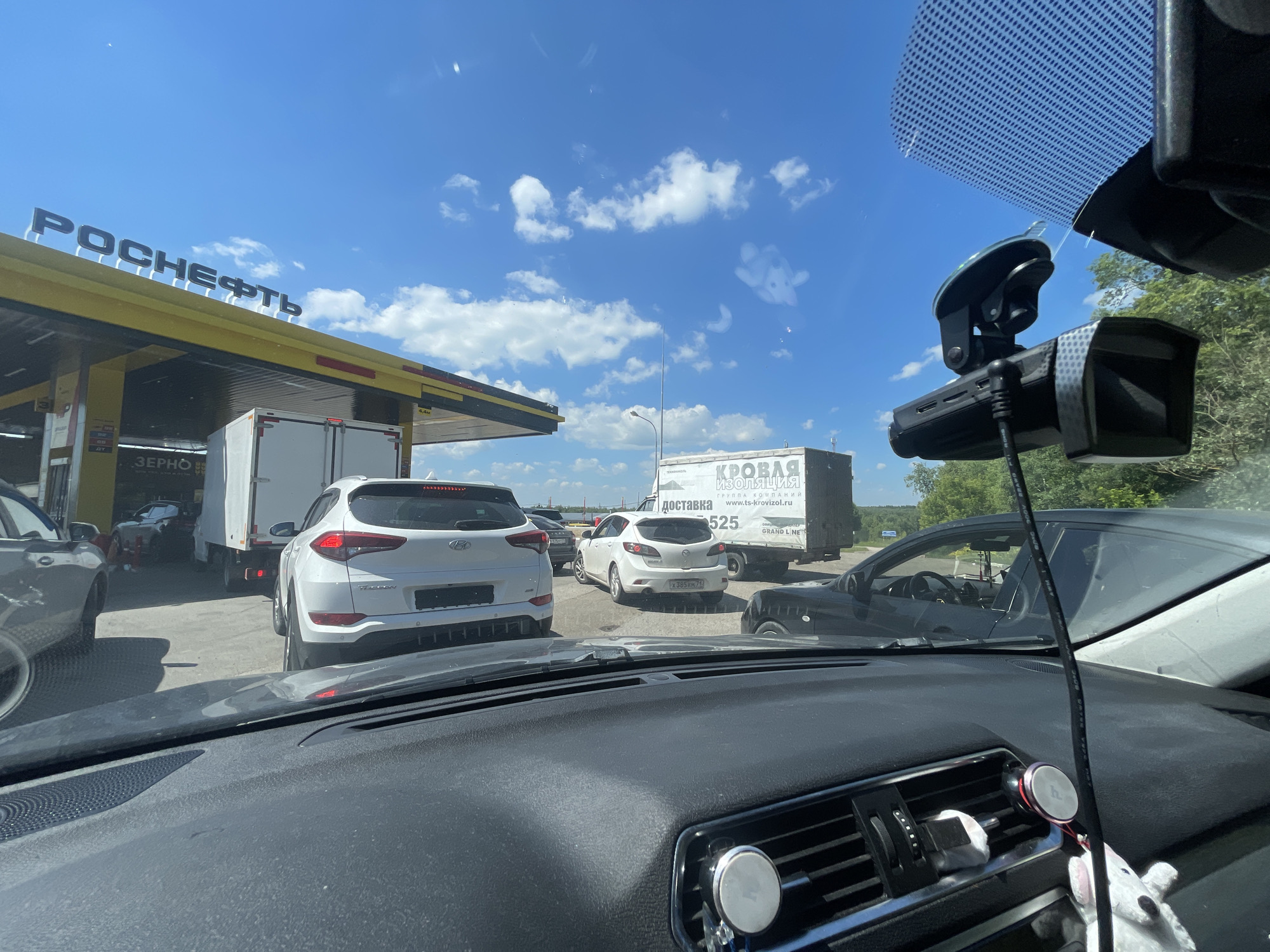 The image depicts a view from inside a vehicle, looking out through the windshield at a traffic jam at a gas station. The gas station canopy has "РОСНЕФТЬ" (Rosneft) signage. Several vehicles are lined up, including a white SUV, a white truck, and other cars. In the background, there are trees and a bright blue sky with some clouds. The dash of the vehicle in the foreground is visible, equipped with a dashcam and a phone mount. There is no visible indication of the specific city, however, the signage "Самылинка, Россия" can be seen on a separate image that is not present in the original image.