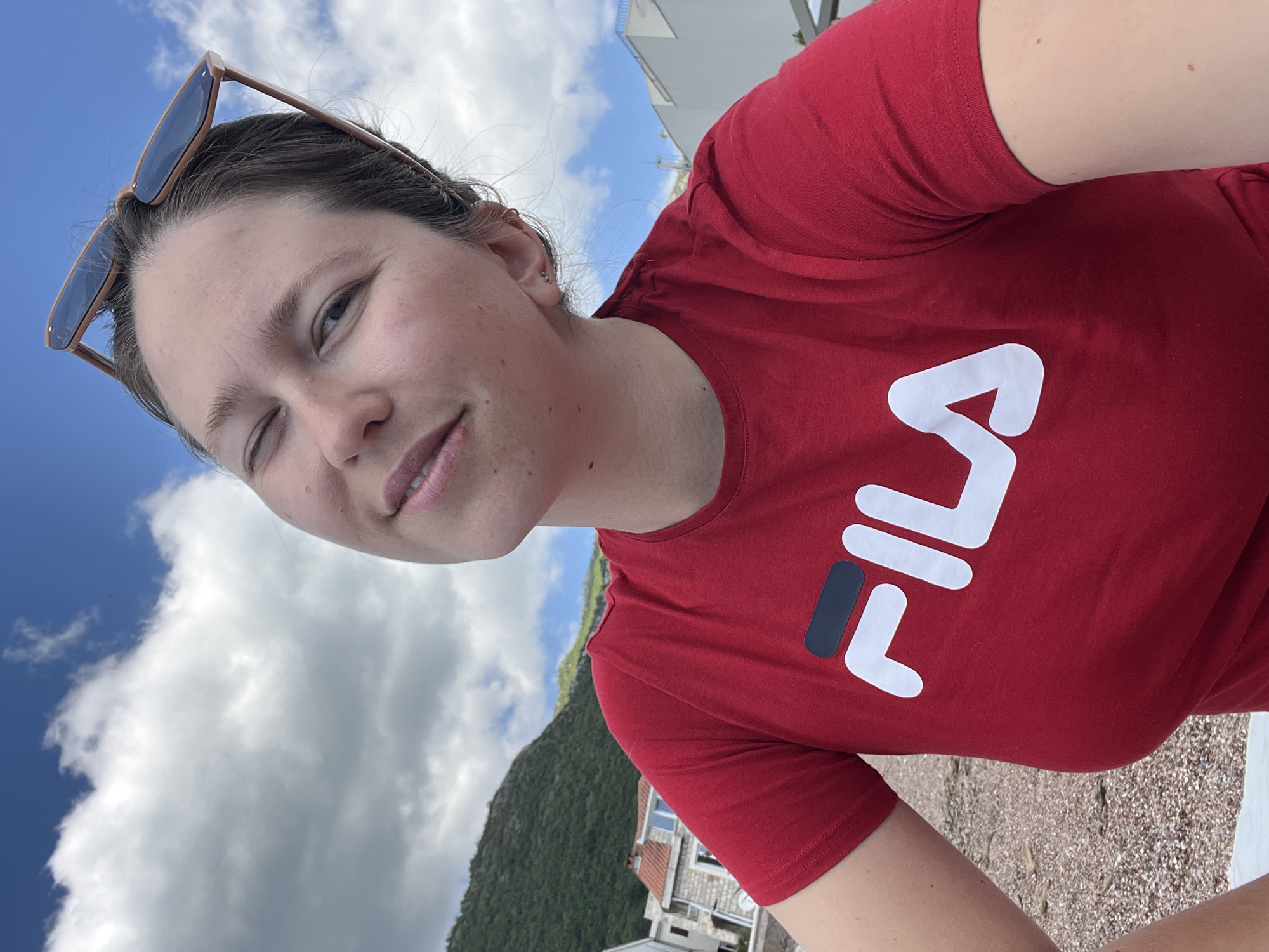 A medium shot depicts a woman with sunglasses on her head wearing a red FILA shirt, winking and looking at the camera. The background suggests an outdoor setting with a cloudy sky above. Below the sky, a green hillside is visible, along with some buildings indicative of the city of Petrovac centar, Montenegro. The ground appears to be a grainy surface, possibly a beach or walkway. The photo seems to be taken at a slight angle.