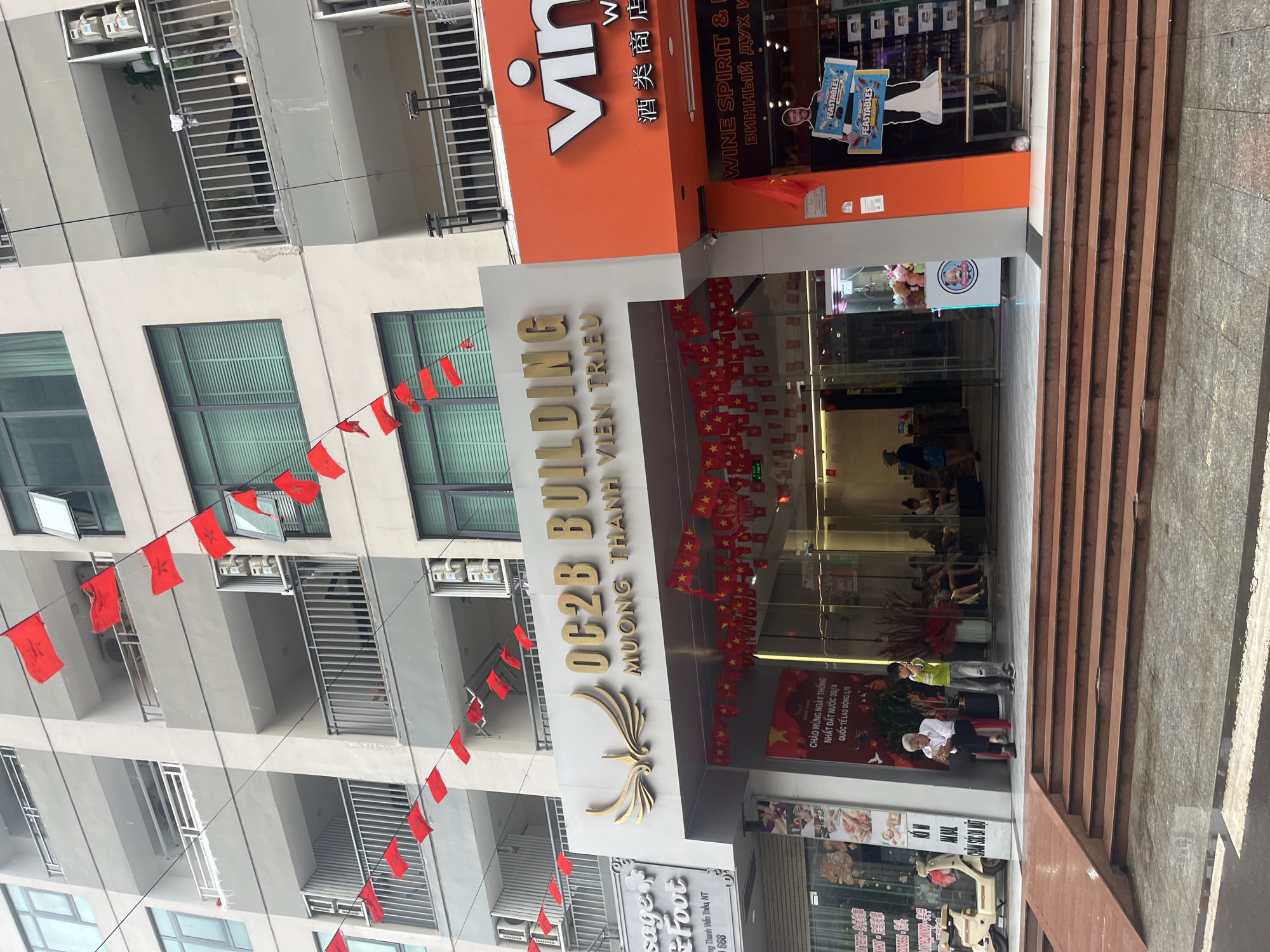 This image, rotated clockwise, depicts the bustling ground level and lower floors of a modern, multi-story building in an urban setting, likely Bac Nha Trang Ward, Vietnam. The architecture features numerous balconies with metal railings and large windows on the upper residential or hotel floors.
The ground floor houses commercial establishments. A prominent sign in gold letters reads "OC2B BUILDING" and "MƯỜNG THANH VIÊN TRIỀU," indicating a key tenant or the building's name, possibly part of the Mường Thanh hotel chain. Another large orange sign for "Vin" (likely VinMart or a related brand) advertises "WINE SPIRIT &," with corresponding text in Chinese ("酒类商店") and Russian ("ВИННЫЙ ДУХ И"). Below this, a storefront features a cardboard cutout of MrBeast promoting "Feastables" products. Further along the facade, a smaller sign mentions "sager Foot" and "ng Thanh Viễn Thều, NT 668," suggesting a massage or spa service.
The building is extensively decorated with strings of red flags, some plain and others featuring a yellow star, resembling the flag of Vietnam. These decorations adorn the exterior, stretching across the facade, and are also visible inside the glass-fronted entrance area. A banner inside the entrance explicitly states "CHÀO MỪNG NGÀY THỐNG NHẤT ĐẤT NƯỚC 30/4 QUỐC TẾ LAO ĐỘNG 1/5," indicating celebrations for National Reunification Day (April 30th) and International Labor Day (May 1st).
Several people are visible near and inside the building's entrance. Some are seated, while others are standing or walking. One individual in a white hat is seated on a red stool near the steps. A short flight of brown steps leads up to the building's main entry on the right side of the frame. The scene appears to be taking place during the daytime, with ample natural light suggesting a clear or partly cloudy day. The overall atmosphere conveys a sense of everyday urban activity mixed with national festive commemoration.
