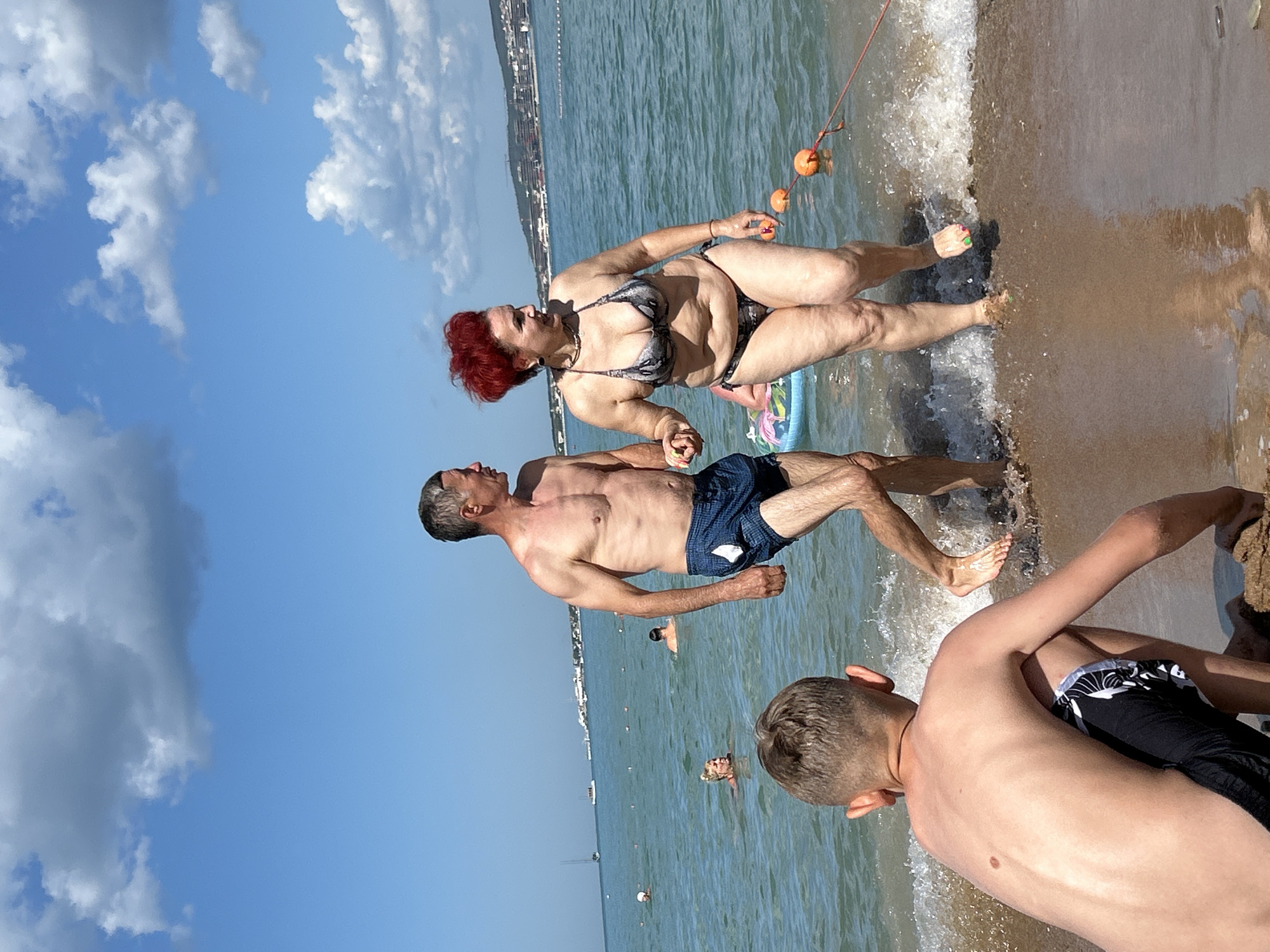 The image depicts a sunny day at the beach in Gelendzhik, Russia. In the foreground, a young boy is seen from behind, kneeling on the sandy shore with waves lapping at his feet. Further out in the shallow water, a middle-aged couple is walking hand-in-hand. The man, wearing blue swim trunks, is looking to his right. The woman, in a patterned bikini, has bright red hair and is also looking to her right. They are both walking towards the shore. In the background, other beachgoers are visible in the water, some swimming and others relaxing. The sea is calm with a few boats sailing in the distance. The sky is blue with scattered white clouds. The coastline is lined with buildings and hills.