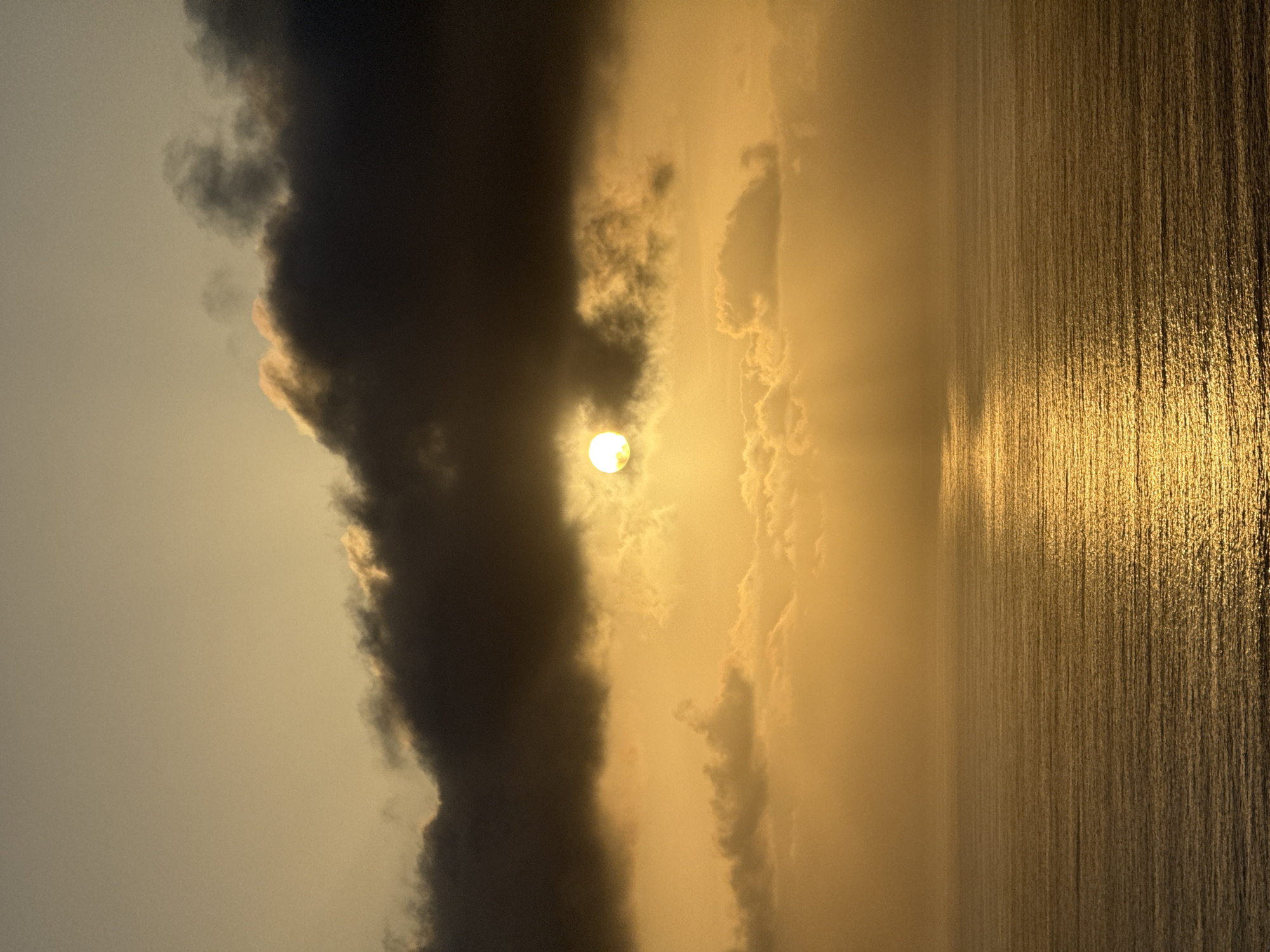 The image depicts a serene ocean scene at sunset in San Diego, California. The sun, a bright orb of light, is partially obscured by dark, wispy clouds, casting a warm, golden glow across the sky and water. The ocean's surface is calm, with gentle ripples reflecting the sunlight, creating a shimmering pathway of light that stretches towards the horizon. The sky is a blend of soft yellows and oranges, with darker clouds adding depth and contrast. The overall atmosphere is tranquil and breathtaking, evoking a sense of peace and natural beauty.