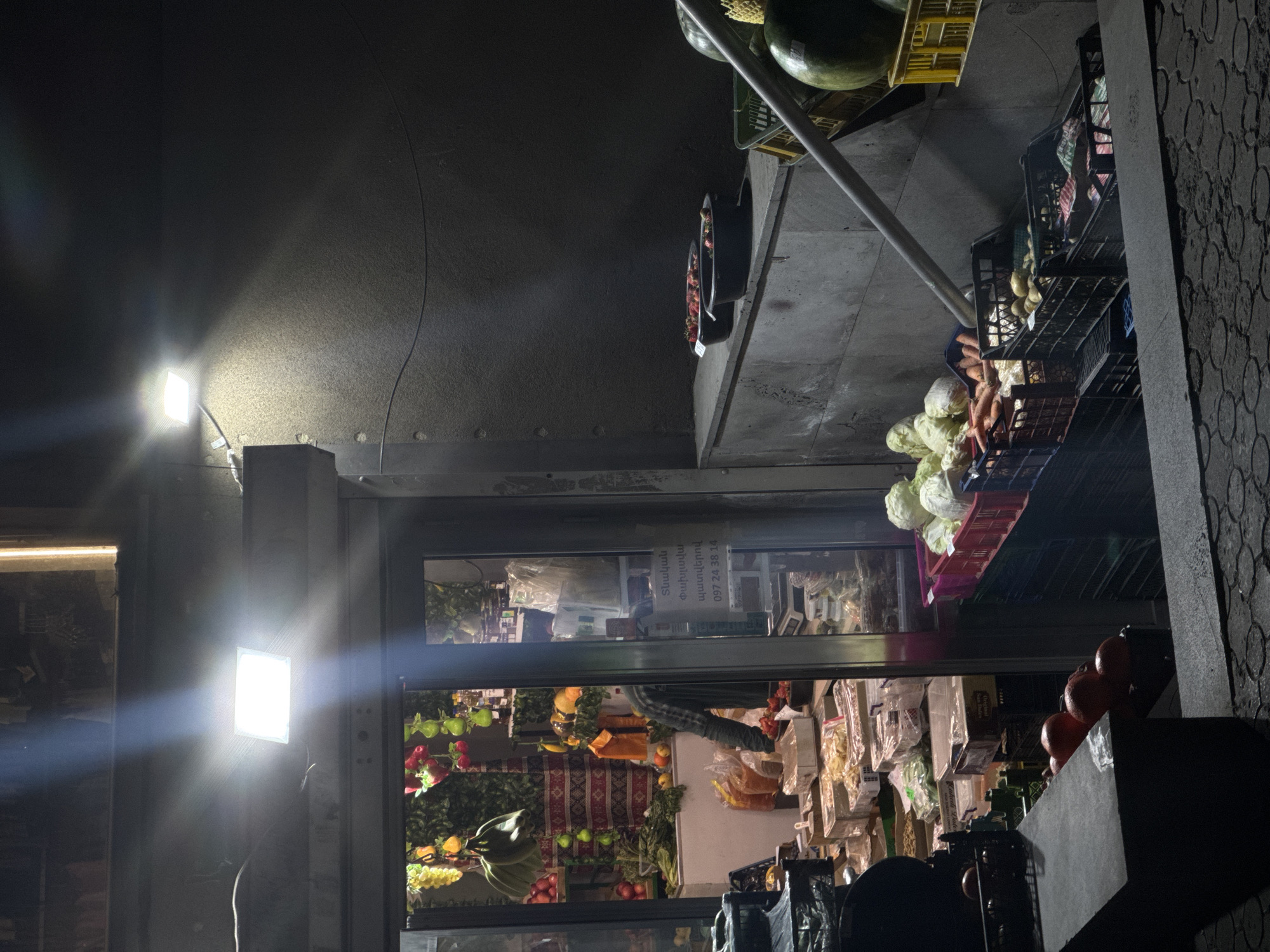 The image shows a nighttime view of an outdoor shop in Yerevan, Armenia, showcasing produce and grocery items. Fruits like watermelons and tomatoes are visible, along with vegetables. There's a small shop window with text that appears to be Armenian writing, indicating it is a storefront. The lighting is uneven, with bright artificial light sources creating glare. The shop is located next to a wall with a patterned surface. The photo's orientation is rotated.