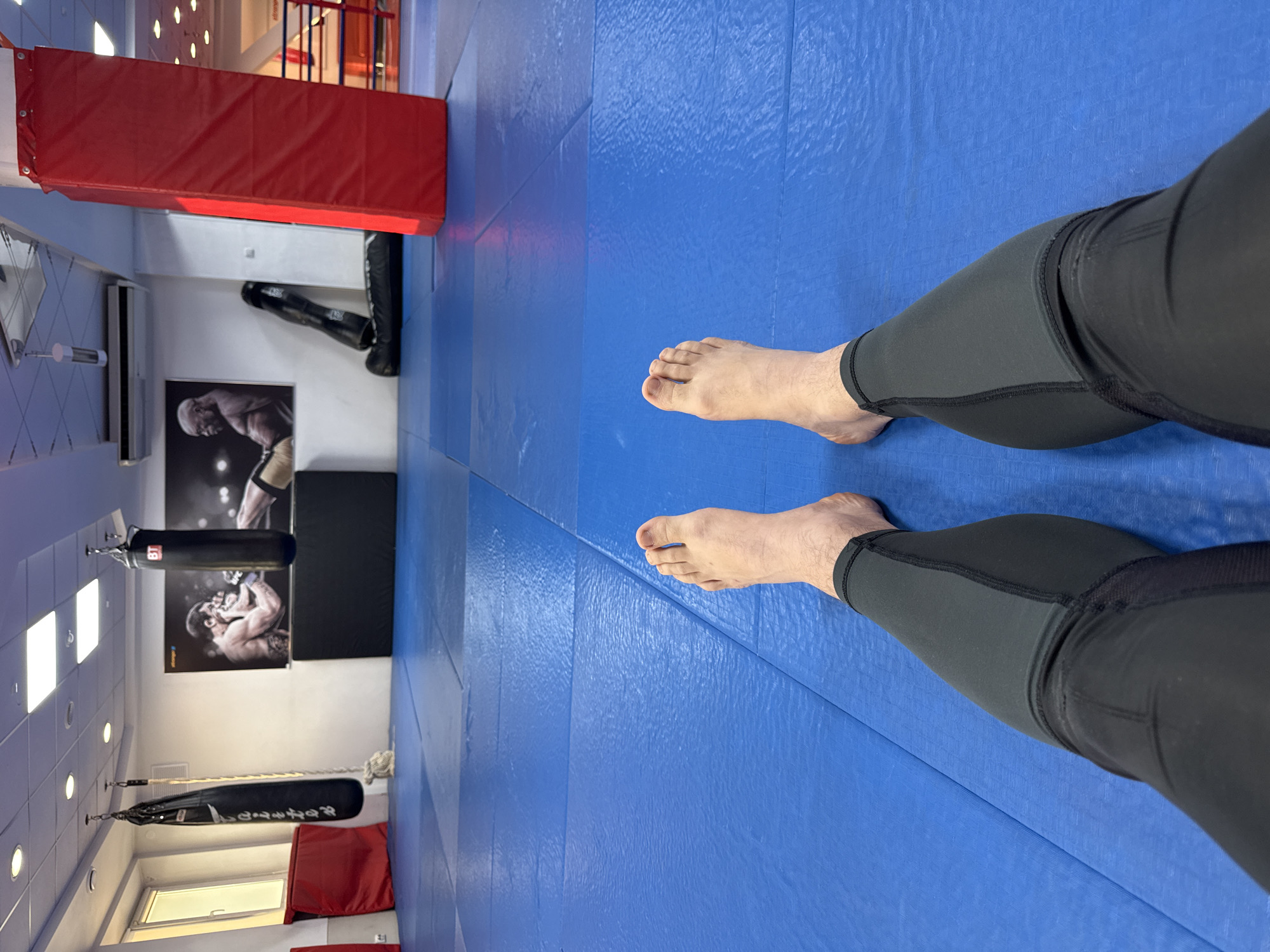 The image shows a pair of legs wearing black compression pants, with bare feet, standing on a blue padded floor in a room with boxing equipment. A red protective wall pad is visible in the background, along with a hanging punching bag and boxing-related posters on the wall. The location is a gym, likely for combat sports training, possibly in the city of Krasnoyarsk, Russia, though there is no definitive evidence to confirm the location other than the user's request to specify it. The person is likely standing still.