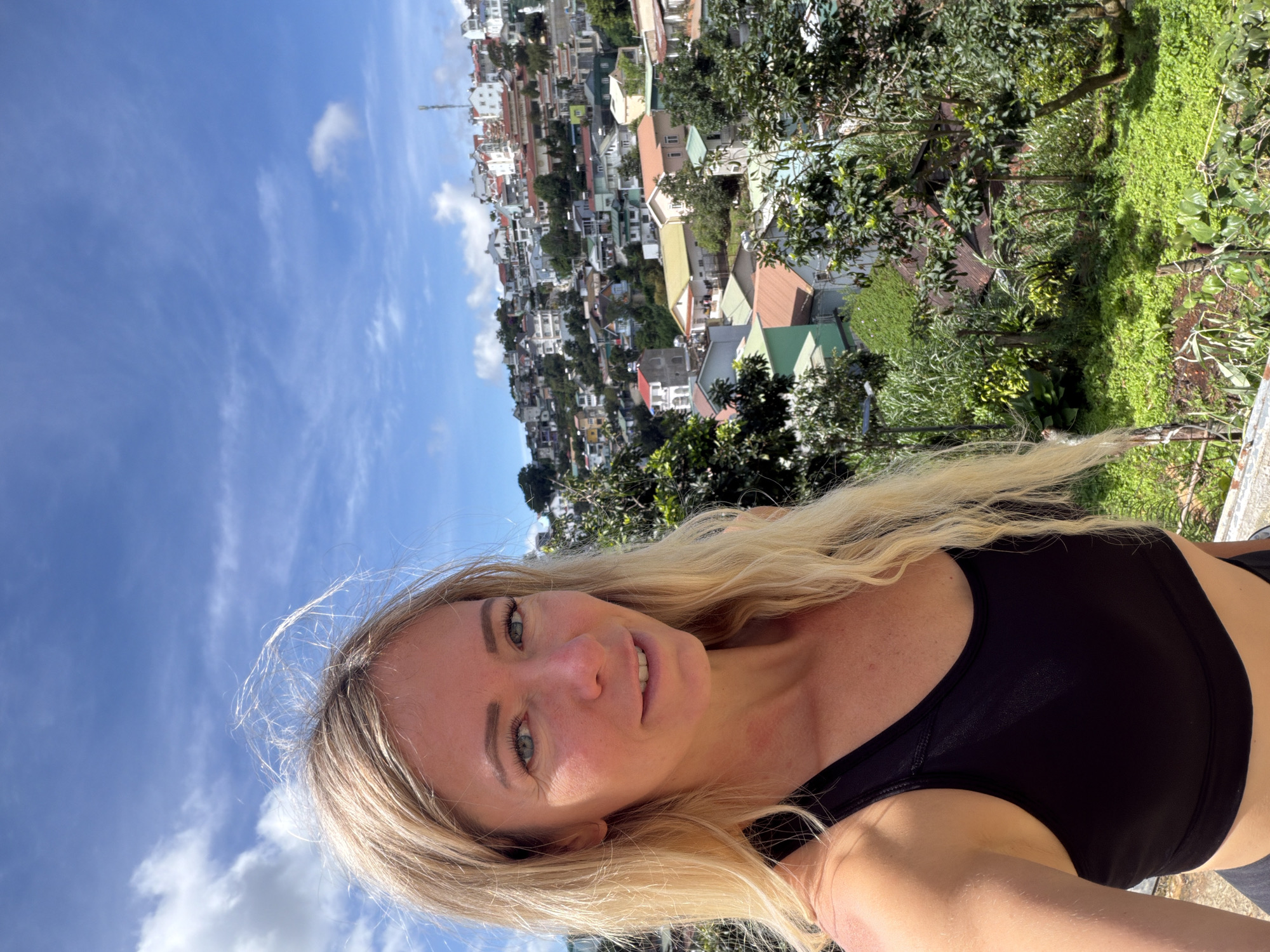 A blonde woman in a black sports bra poses for a selfie outdoors, with a picturesque view of Da Lat, Vietnam, in the background. The city, nestled in a hilly landscape, is characterized by its colorful houses with red and orange tile roofs, interspersed with lush green trees and vegetation. The sky is a vibrant blue with scattered white clouds, suggesting a bright, sunny day. The woman's hair is windblown, adding a dynamic element to the portrait. The foreground shows her upper body and a glimpse of her lower torso, while the middle ground is dominated by the densely packed houses and trees of Da Lat. The overall impression is one of a traveler enjoying the scenic beauty of the city.