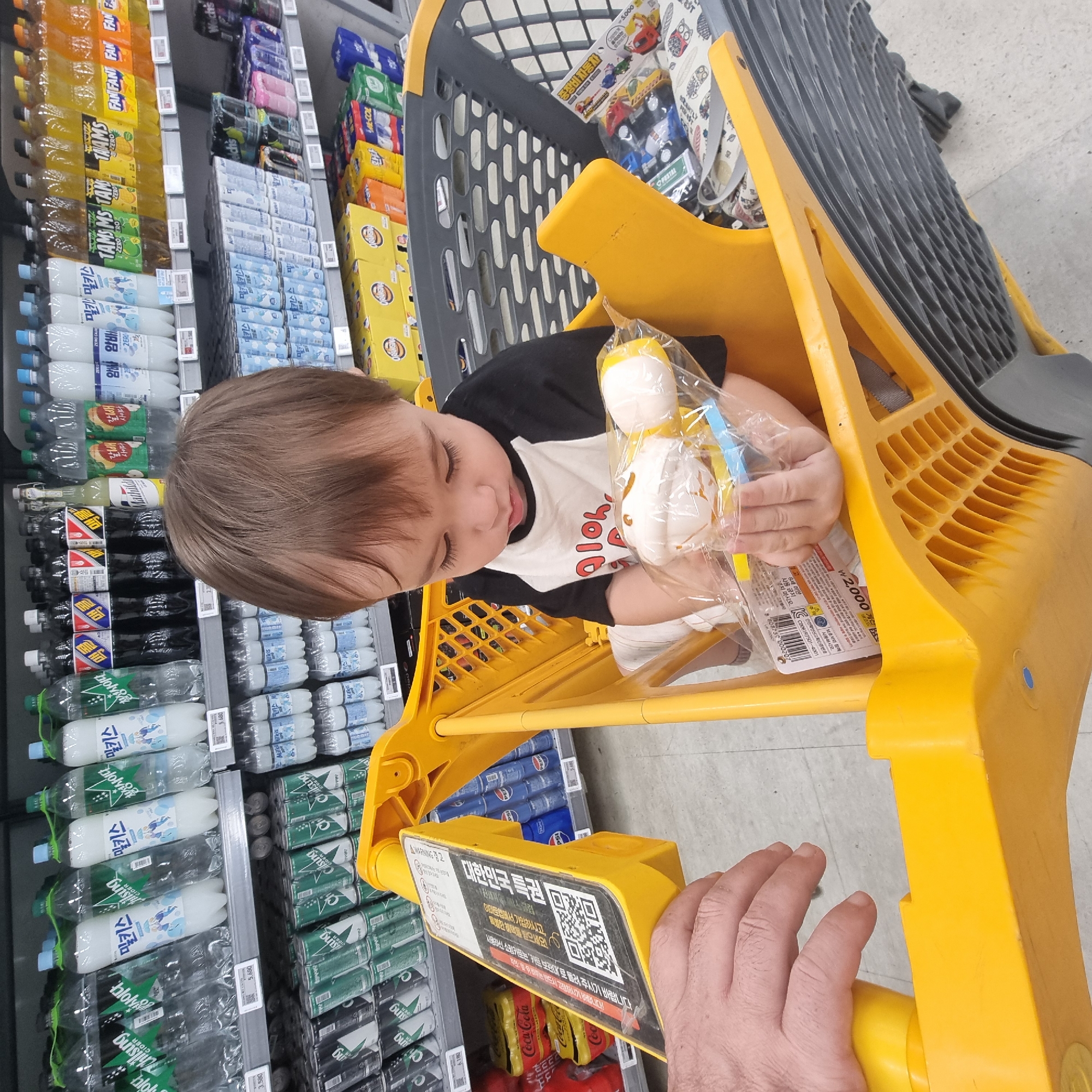 The image shows a young child, likely a toddler, sitting in a yellow shopping cart in a grocery store aisle. The child is wearing a black and white raglan t-shirt with a graphic design. The child is holding a toy that is partially visible inside a clear plastic bag. The shopping cart is filled with various items in the background, including bottled drinks on shelves, cans, and boxes of goods. A person's hand is visible on the left side of the image, gripping the handle of the shopping cart. The environment appears to be a well-lit supermarket. Based on the product packaging and price tags in Korean, this scene is taking place in Wonju-si, South Korea.