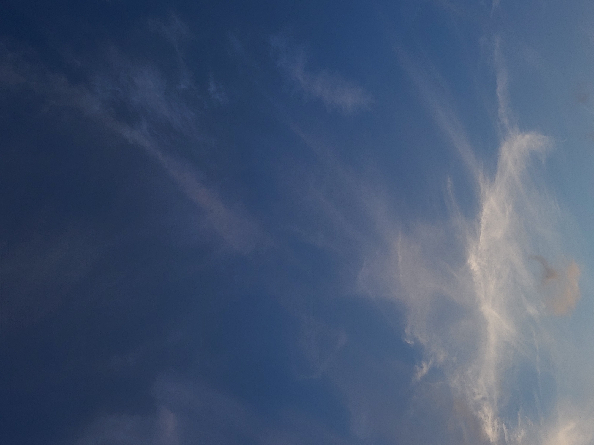 The image shows a vast expanse of a deep blue sky with wispy, thin clouds scattered throughout. The clouds are white and appear to be cirrus clouds, streaking horizontally across the frame. Some areas of the sky are clear and deep blue, while others are more diffused by the faint cloud cover. Towards the bottom of the image, the clouds become slightly denser and more prominent, with hints of lighter blue and white. There are no discernible objects or people in the image, and the scene is solely focused on the atmospheric conditions of the sky. The lighting suggests it could be daytime, possibly late afternoon or early evening given the depth of the blue. The location is unknown.