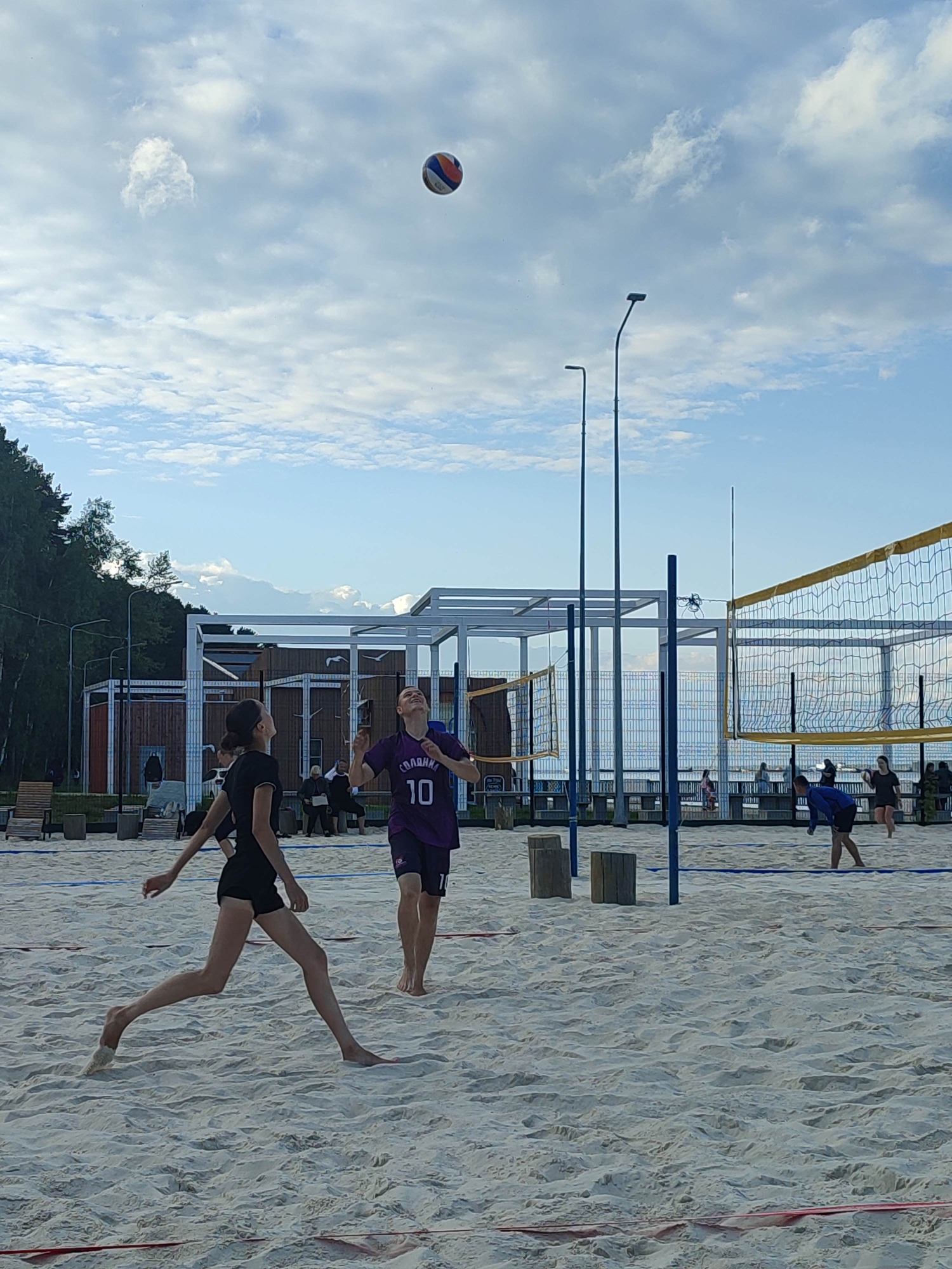 This media file shows a beach volleyball game in progress. The scene is set on a sandy beach with a volleyball net and court clearly visible. In the foreground, two young women are actively playing. One, in a black top and shorts, is in motion, her body angled as if reaching for or hitting the ball. The other player, wearing a purple jersey with the number "10" and dark shorts, is looking up at the ball, which is in the air above them.
In the background, several other people are visible, some spectating and others appearing to be involved in a game on another court. There are structures resembling beachside pavilions or buildings behind the courts, and trees line the background, suggesting a coastal or lakeside setting. The sky is blue with scattered, fluffy white clouds, indicating daytime with potentially pleasant weather. The lighting suggests it could be late afternoon or early evening.
The visible text on the purple jersey reads "СОЛДАНКА" and the number "10". The overall atmosphere appears to be one of active recreation and sport.