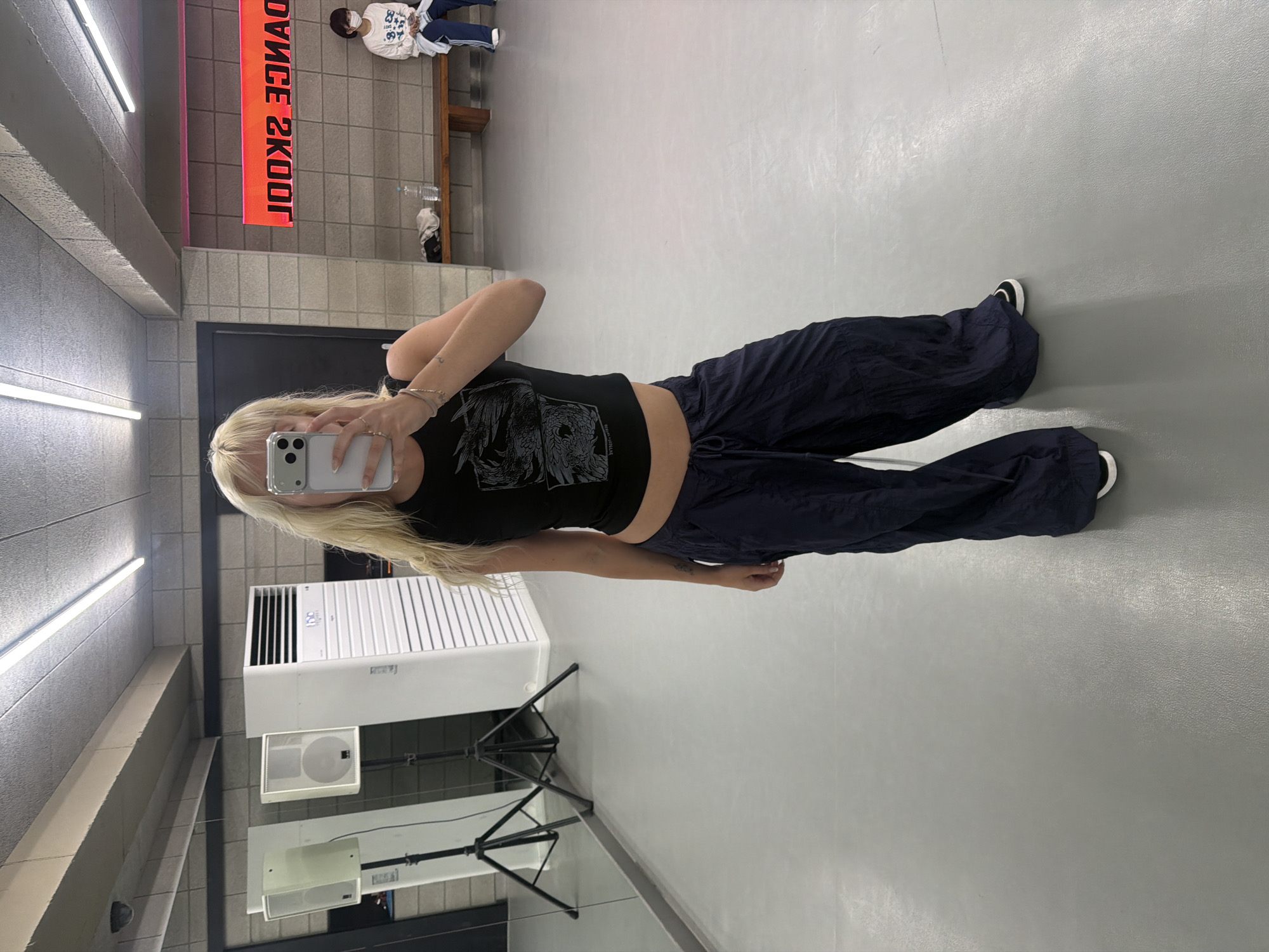 The image shows a woman standing in a dance studio. She is holding her phone up to take a selfie in the mirror. She is wearing a black crop top and dark blue parachute pants, with black and white shoes. Behind her, there is an air conditioning unit and a mirror on the wall. The studio has a gray floor and a brick wall with a sign that says "DANCE SKOOL" in red letters. In the background, two figures are seen, one sitting on a bench. The location is likely in Seoul, South Korea, given the Korean characters on the sign and the general appearance of the environment.