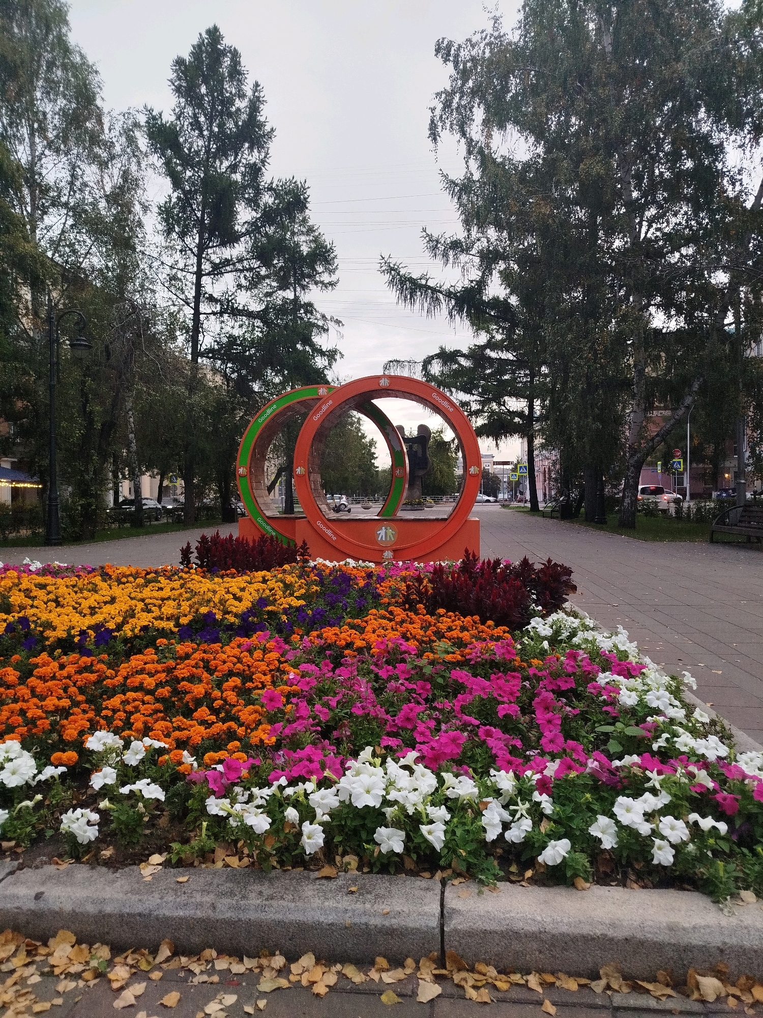 The image depicts a park in Kemerovo, Russia, specifically on Sovetsky Prospekt. In the foreground, a vibrant flowerbed overflows with an array of colorful blooms, predominantly orange, yellow, pink, and white petunias, with some purple flowers interspersed. The flowerbed is bordered by a concrete curb, and fallen autumn leaves are scattered on the gray pavement. Behind the flowerbed, an abstract orange sculpture featuring two interconnected rings stands prominently. A dark silhouette of a statue is visible within the rings. The background reveals a tree-lined walkway with benches and lampposts. Beyond the trees, buildings and cars suggest an urban setting. The sky is overcast, indicating a cloudy day.