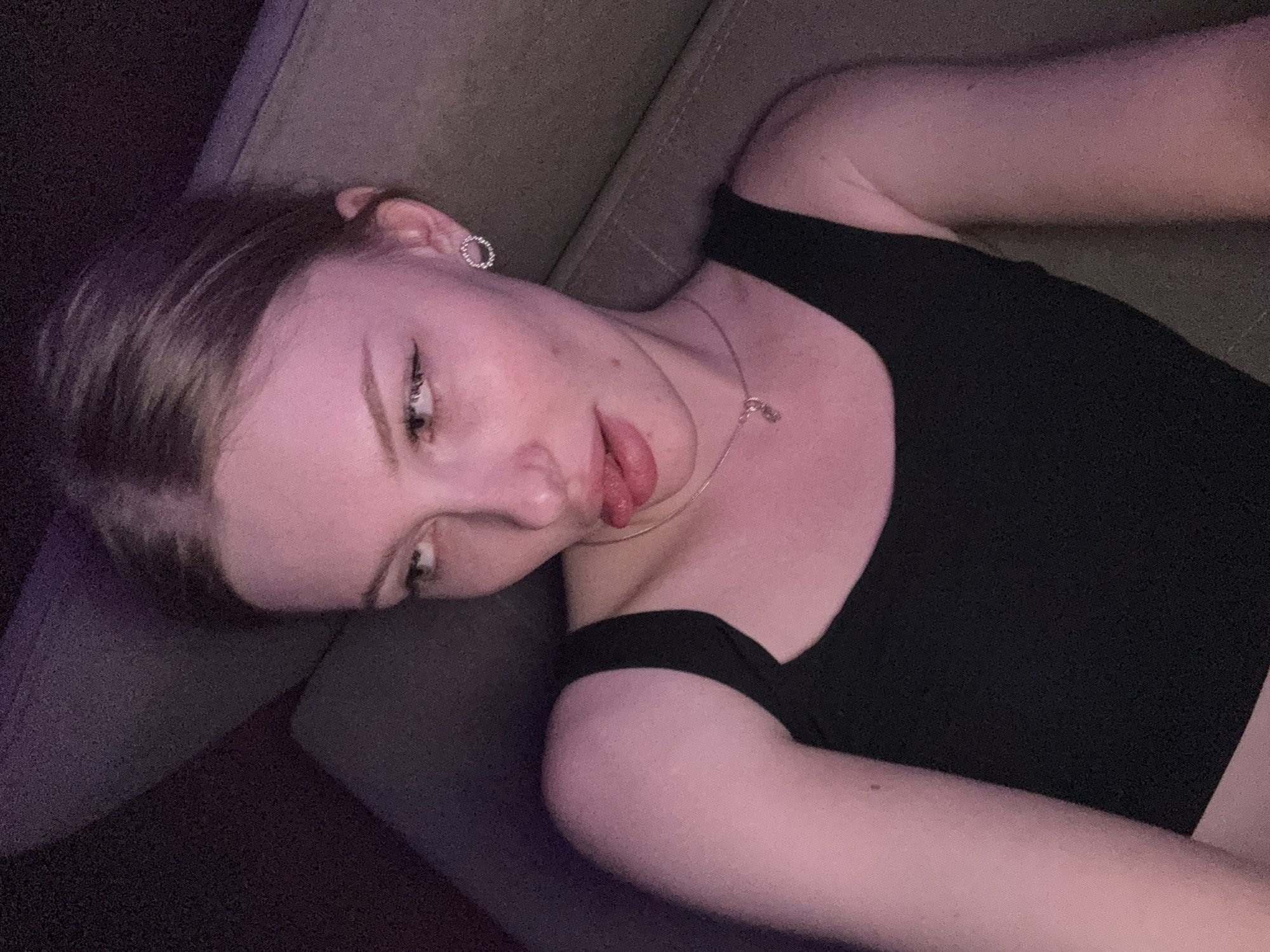 A young woman with fair skin and blonde hair lies on a taupe-colored upholstered surface, possibly a couch. She wears a black off-the-shoulder top and a delicate necklace. Her left ear is adorned with a small, round earring. Her head is tilted towards the left, with her mouth slightly open and tongue protruding. The lighting is dim, with a purplish hue casting shadows, particularly noticeable on the couch in the lower left corner. The scene is indoors. There is no evidence of the city of Perm, Russia.