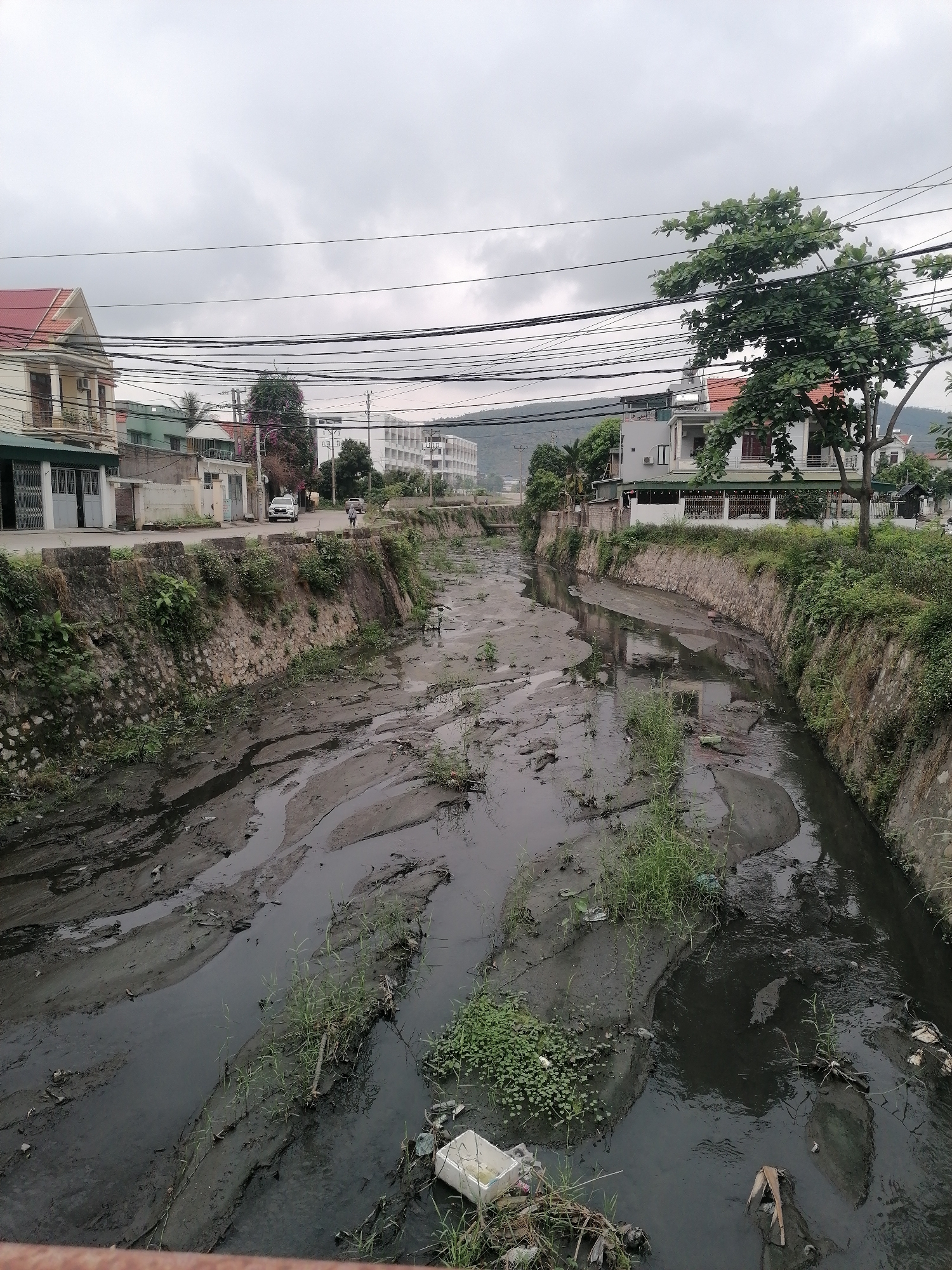 The image depicts a stagnant, polluted canal or riverbed located in Ha Tu Ward, Vietnam. The water is dark and murky with visible debris and litter scattered throughout. The canal banks are lined with stone or concrete walls, with overgrown vegetation and weeds sprouting from the edges. In the background, there are residential buildings and a multi-story building. Overhead, a dense network of utility wires stretches across the sky. Two people are walking in the background on the left side of the frame. A car is parked on the street to the left of the frame. The sky is overcast, suggesting a cloudy day.