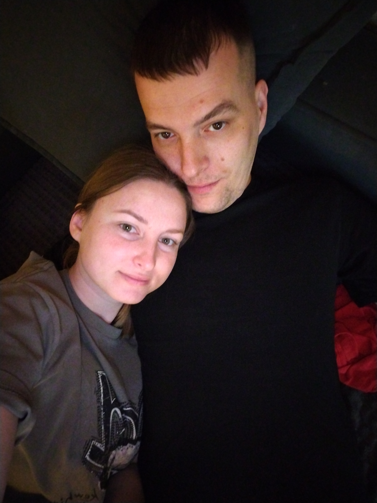 A photo depicts a man and a woman posing for the camera. The man has short dark hair and is wearing a black shirt. The woman has light brown hair and is wearing a gray t-shirt with a graphic design. The background is dark, suggesting an indoor setting, likely a home. Red fabric is visible in the background. The exact location in Chaikovsky, Russia, cannot be determined from the image.