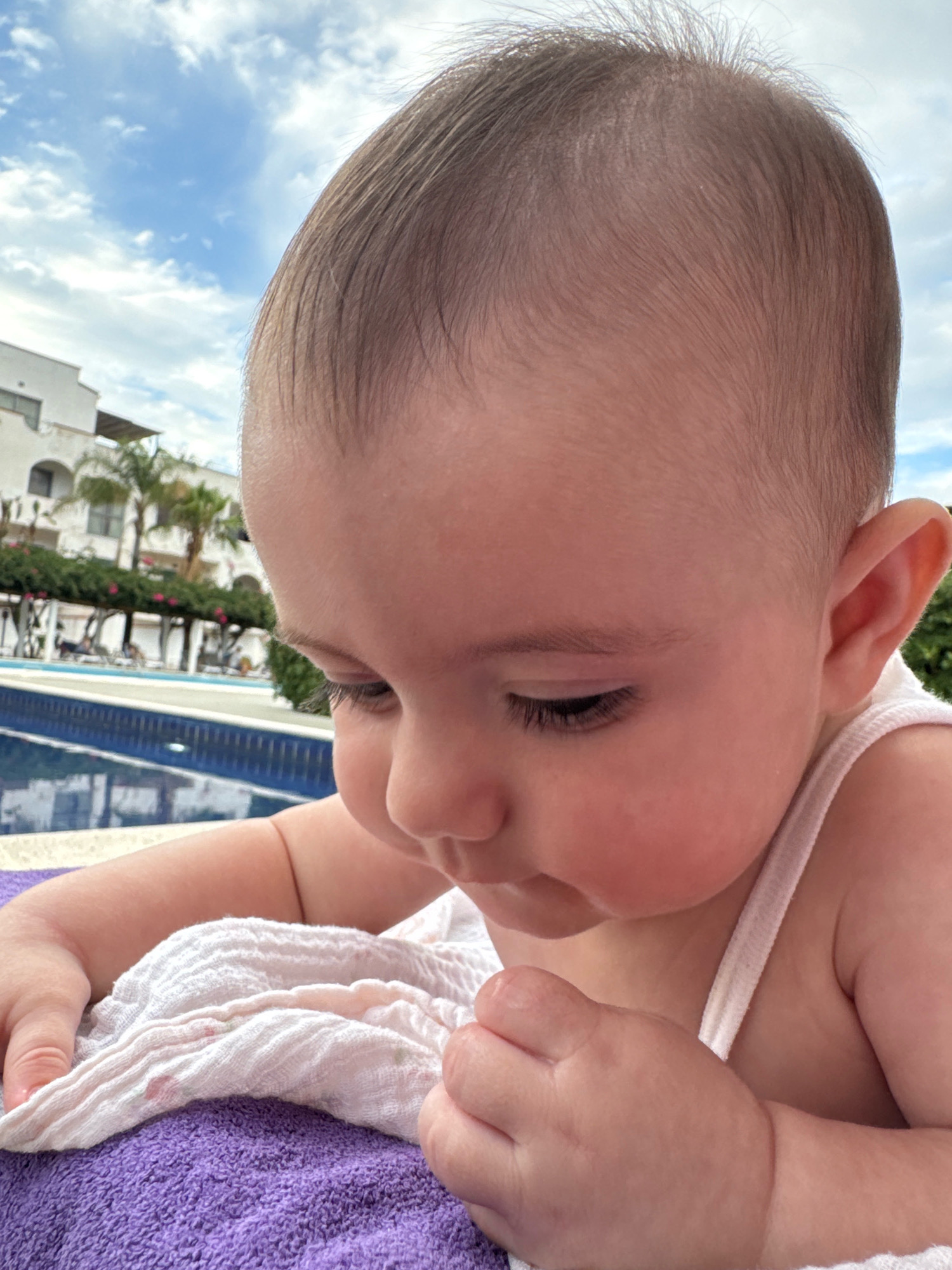 A close-up, head-and-shoulders shot of a baby looking down, with their head angled to the right. The baby has fair skin and fine, light brown hair. They are wearing a white tank top, and their hands are resting on a white, crinkled cloth. The cloth is on top of a purple towel. In the background, out of focus, is a swimming pool with blue tiles and a reflection of the sky. Beyond the pool are buildings, palm trees, and a cloudy blue sky. The scene appears to be outdoors, possibly at a hotel or resort in Isola delle Femmine, Italy, given the architectural style and vegetation.
