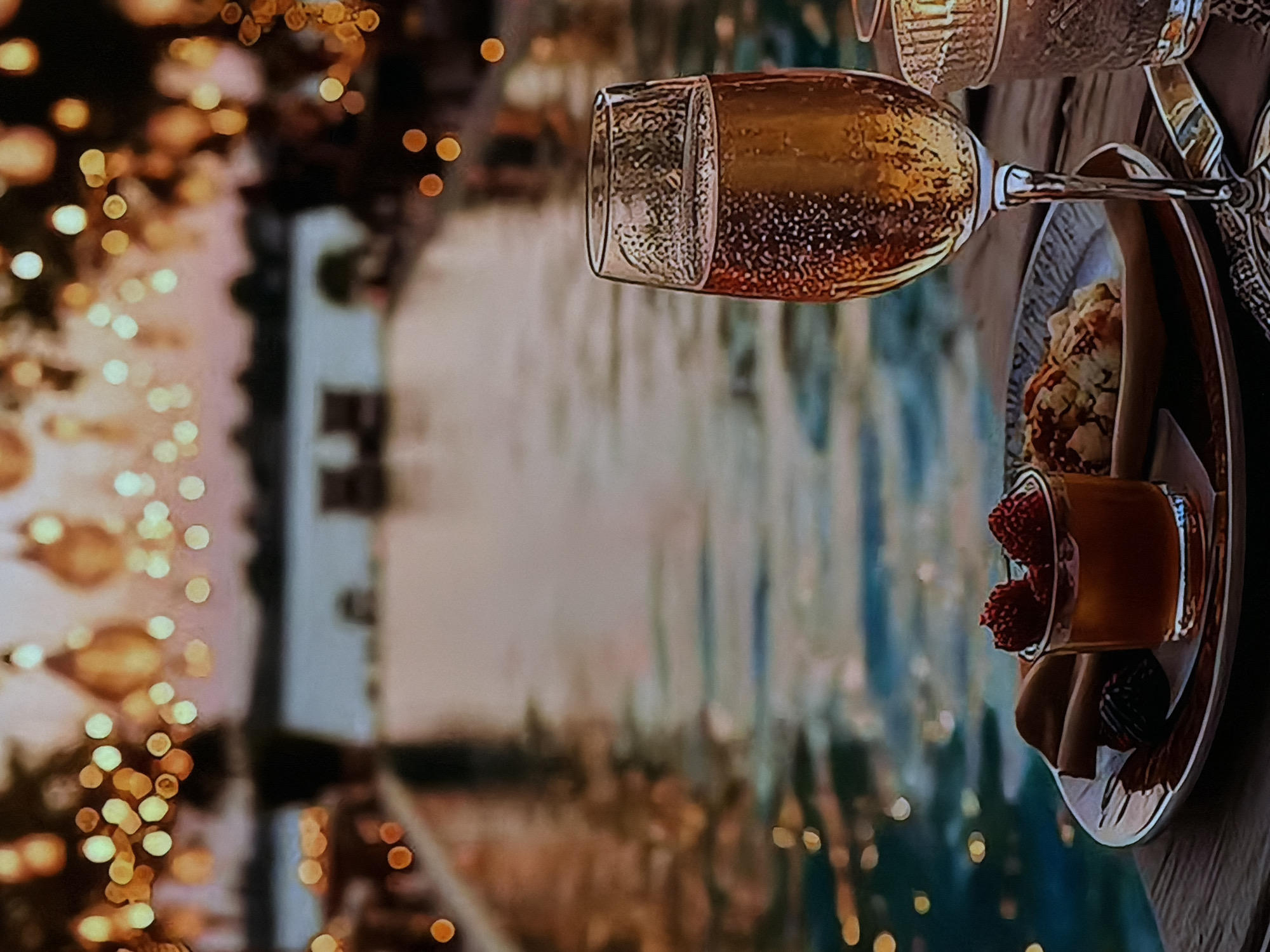 An evening scene captures a glass of sparkling wine, likely champagne or prosecco, bubbling and positioned to the right of the frame. The glass, reflecting warm light, is held by a stem that angles towards a small dessert plate. On the plate, there are several confections: a small, intricately textured pastry or tart, a shot glass or ramekin filled with bright red raspberries and a viscous liquid (possibly a sauce or liqueur), and a rolled dessert that resembles a waffle or crepe. Another glass, partially visible, sits further back on the right.
The background is softly blurred with numerous golden and white bokeh lights, suggesting fairy lights or distant city lights, creating a festive and romantic ambiance. Behind the foreground elements, a tranquil body of water, possibly a swimming pool or a canal, reflects the ambient light, appearing calm and serene. Faint outlines of structures or buildings are discernible across the water, adding depth to the scene. The overall impression is one of a relaxed, luxurious, and possibly celebratory occasion taking place outdoors at dusk or night, perhaps at a resort or an upscale venue in Sofiivska Borshchahivka, Ukraine. There is no visible text in the image.