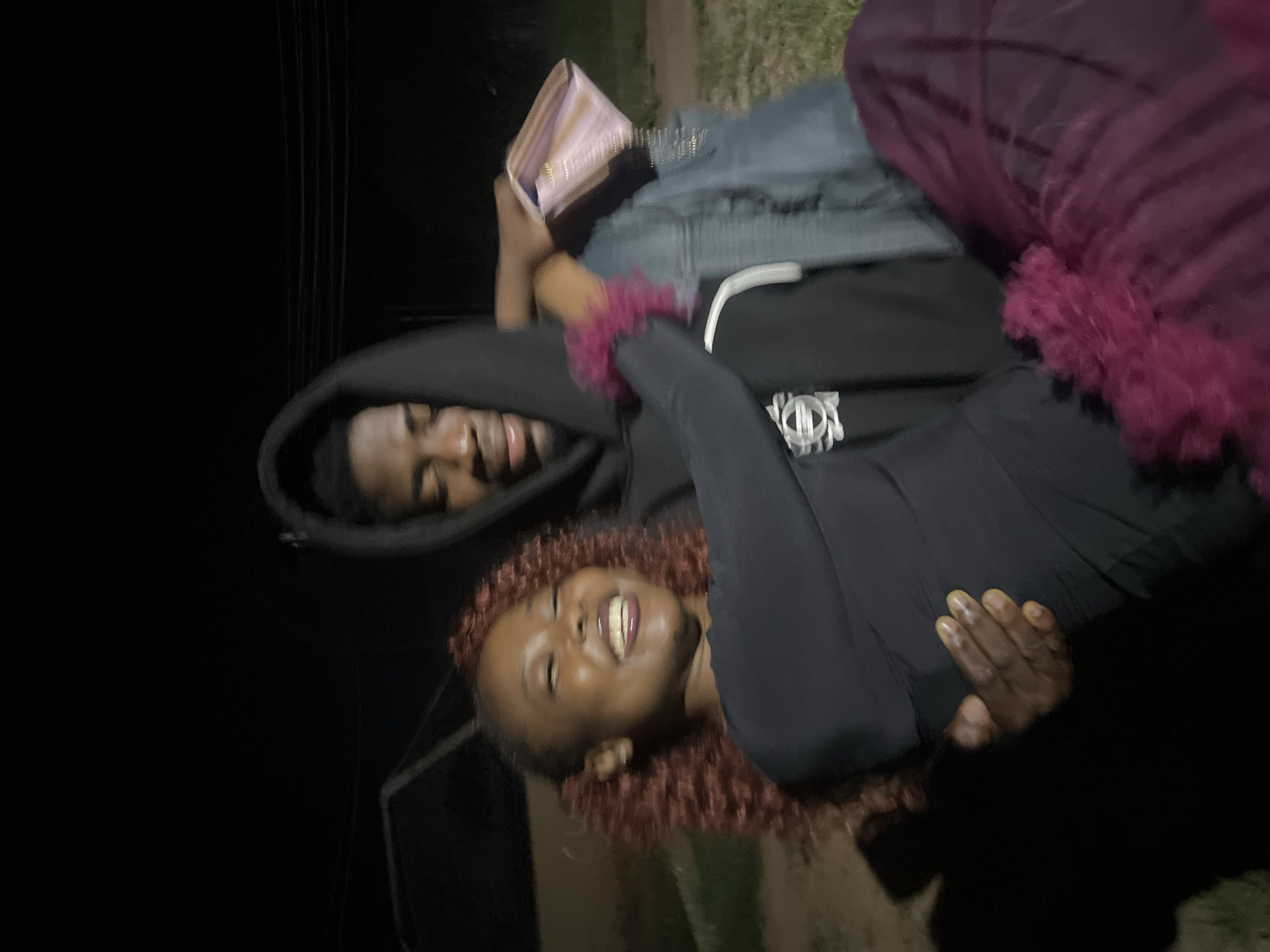 In an outdoor, nighttime scene in Durumi, Nigeria, a young man carries a young woman in his arms. The woman, with curly reddish-brown hair styled into two pigtails, is laughing heartily and looking to her left. She wears a black long-sleeved top and a dark purple skirt with a fluffy trim. The man, wearing a black hoodie, is looking down at the woman and holding her securely. He is carrying a light pink clutch purse in his right hand, and she is holding onto his arm with her left hand. The background is dark, with some faint outlines of structures and overhead wires visible. The ground appears to be covered in grass and dirt. The overall impression is one of affection and joy.