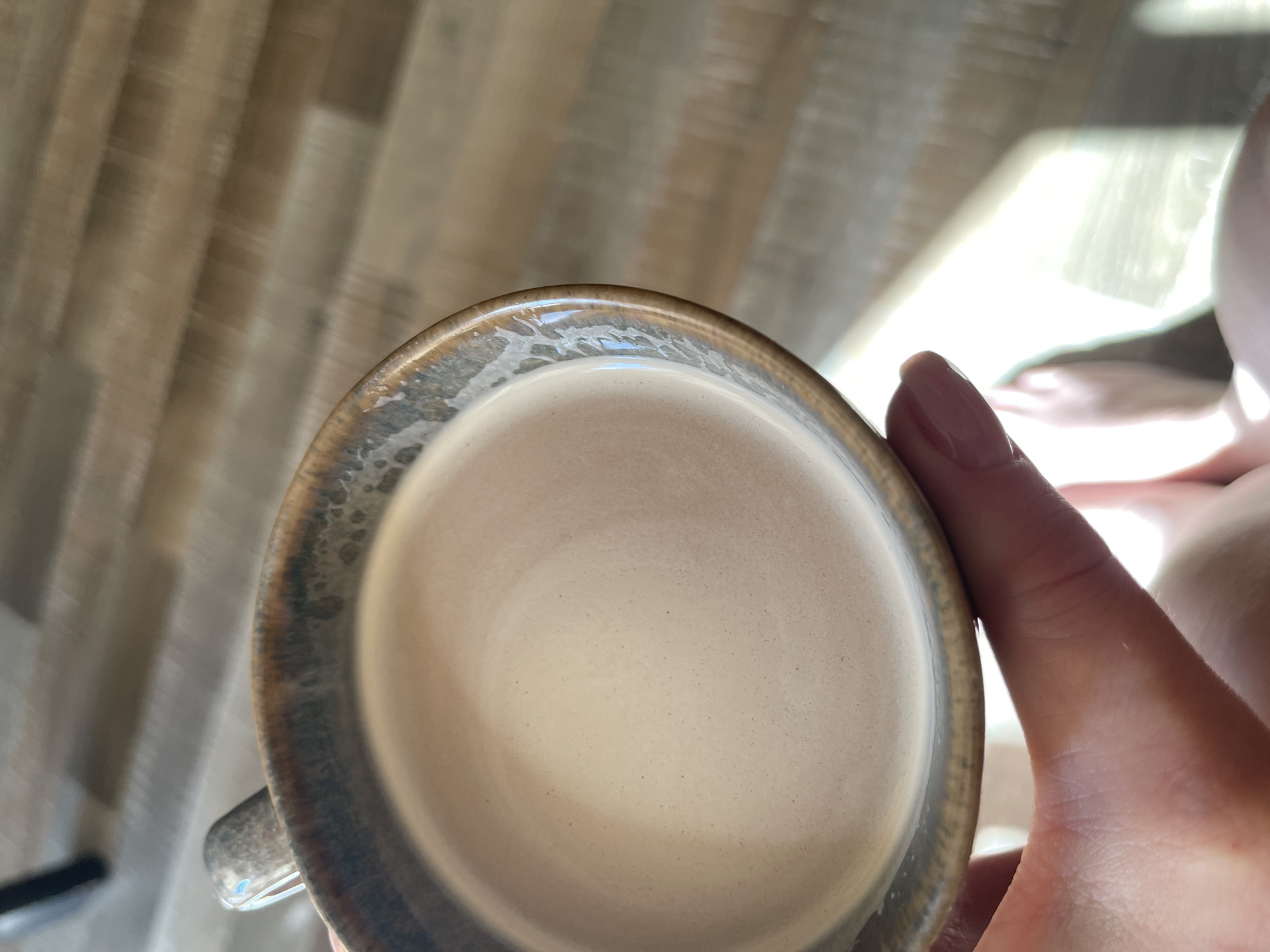 A close-up shot shows a person holding a ceramic mug filled with a light-brown liquid, possibly coffee or tea, with a frothy top. The mug has a speckled, glossy finish along the rim. The background consists of a wooden floor with visible planks, bathed in sunlight that creates distinct light and shadow patterns. A person's bare feet are visible in the lower right corner, suggesting the scene takes place indoors, likely in a home or cafe. The location of Kommunarka, Russia is not discernible from the visual information provided.