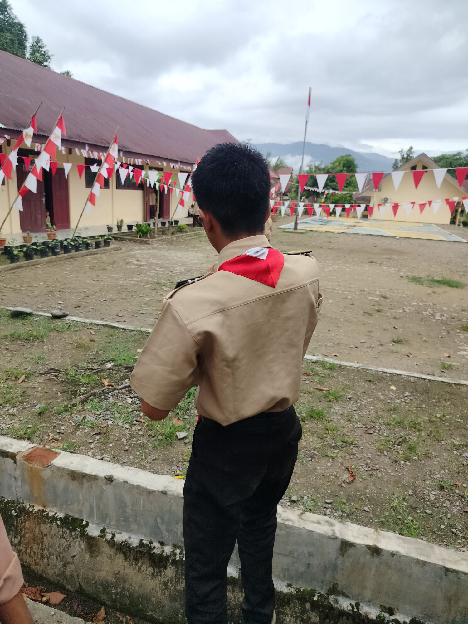 The image depicts a young male scout, viewed from behind, standing in what appears to be a schoolyard in Purwodadi, Indonesia. The scout is wearing a tan uniform shirt with red and white insignia on the collar and dark pants. Behind him, a building with a reddish-brown roof is visible, adorned with red and white triangular flags and Indonesian flags. The ground is a mix of dirt and gravel, with some patches of grass and scattered leaves. In the background, more buildings decorated with flags and a flagpole with the Indonesian flag can be seen. The sky is overcast with clouds. The overall scene suggests an outdoor event or ceremony taking place at an educational institution.