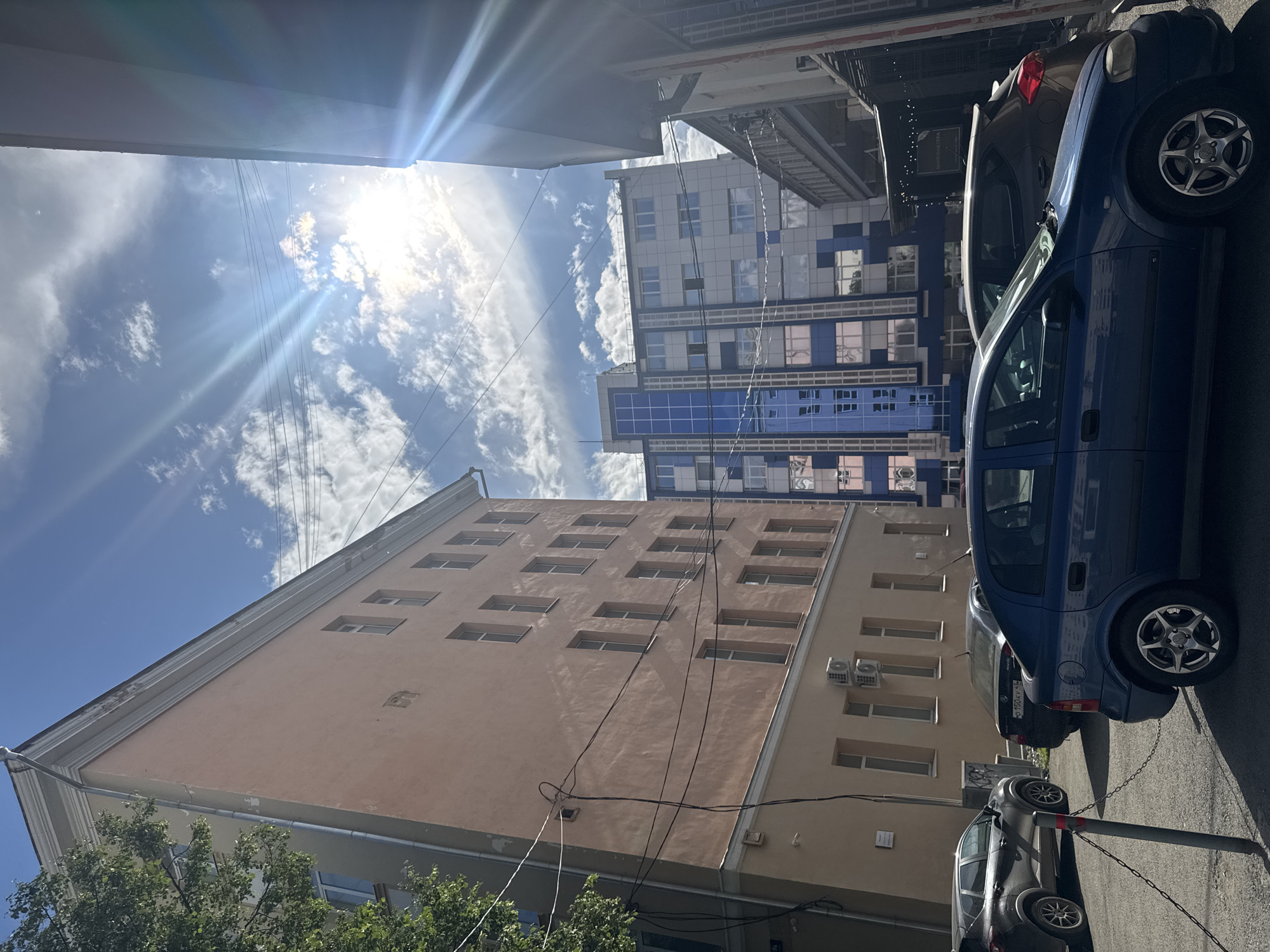 The image shows a street scene in Yekaterinburg, Russia. The perspective is looking upwards towards the sky and buildings. The sun is bright and there are some clouds in the sky, suggesting it is daytime and the weather is fair.
The main subjects are buildings and cars. There are several buildings visible. One building in the foreground is tall and peach-colored with many rectangular windows arranged in rows. To the right, a modern building with blue and white panels and many windows is visible. Further in the background, another building is partially visible.
Parked on the street are three cars. A dark gray car is parked closest to the camera, followed by a blue car, and then a darker car partially obscured. The cars are parked in a row on the asphalt road.
There are some electrical wires running across the scene, strung between buildings. Visible on the dark gray car is a license plate with Russian characters. A sign with text is visible on the dark car in the background, though the text is not clearly legible in the original image.
The overall scene depicts an urban environment on a sunny day. The upward angle and the presence of buildings and cars are typical of a city street.