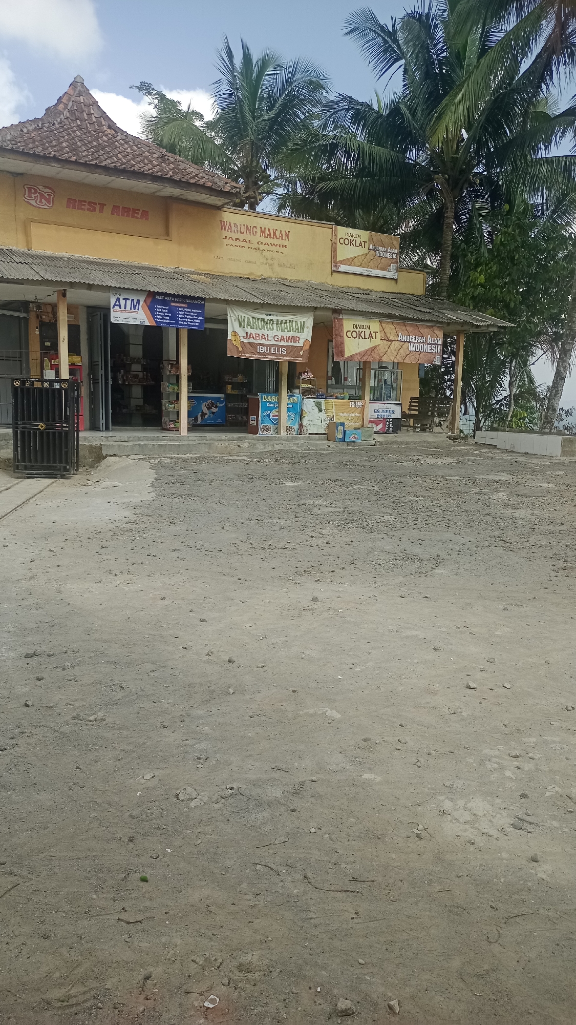 The image depicts a storefront with a sign that reads "PN REST AREA" on the left side. Next to it, a sign for "WARUNG MAKAN JABAL GAWR" is visible. The building is painted yellow and has a tiled roof. Several banners are hanging from the storefront, including one that says "WARUNG MAKAN JABAL GAWR IBU ELIS." There are also signs for "DIARUM COKLAT ANUGERAH ALAM INDONESIA."
The storefront has an open entrance, revealing shelves stocked with goods. An ATM machine is also visible to the left of the entrance. A black metal gate stands in front of the entrance. The ground in front of the building is covered with gravel. Palm trees are visible behind the building. The scene appears to be in Karyamandala, Indonesia, based on the text on the signs.