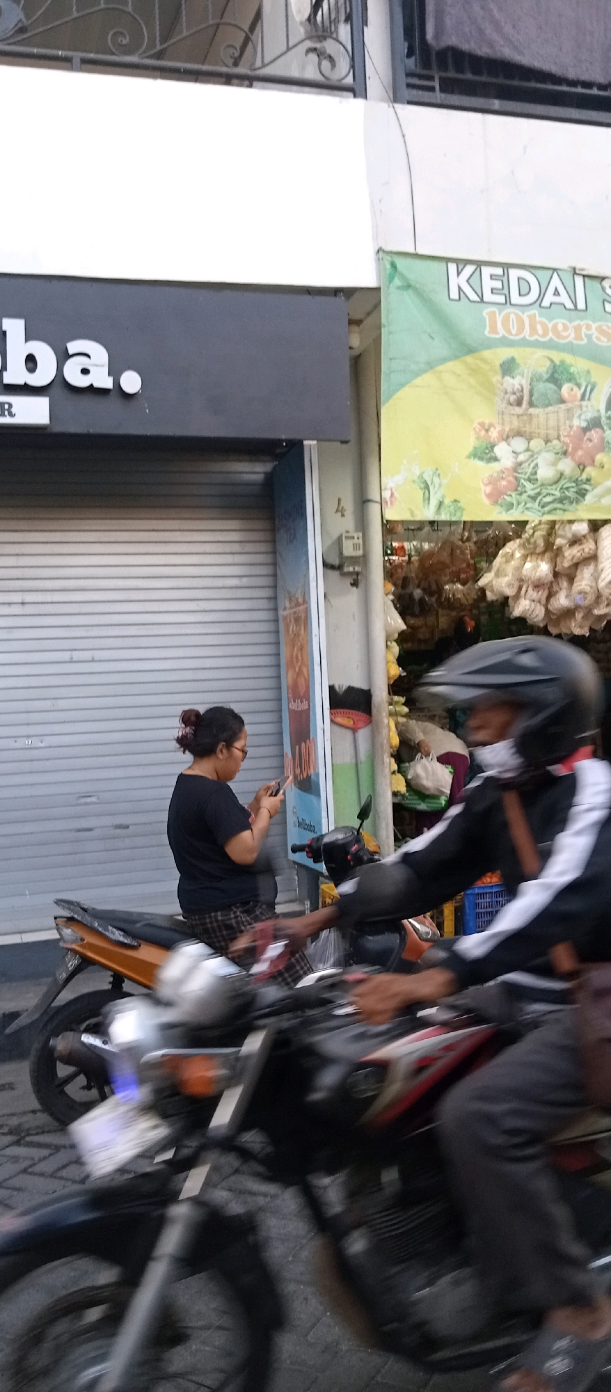 The image shows a street scene in Wadungasri, Indonesia. A woman with her hair in a bun is sitting on a motorcycle, looking at her phone. She is wearing a black t-shirt and plaid pants. In the background, there is a store with a closed metal gate and a sign that reads "boba." To the right, a green banner advertises "KEDAI SERBA 10rb" and shows an illustration of vegetables. Another person on a motorcycle, wearing a black helmet and mask, is in the foreground, slightly blurred due to motion. The street is paved with interlocking bricks.