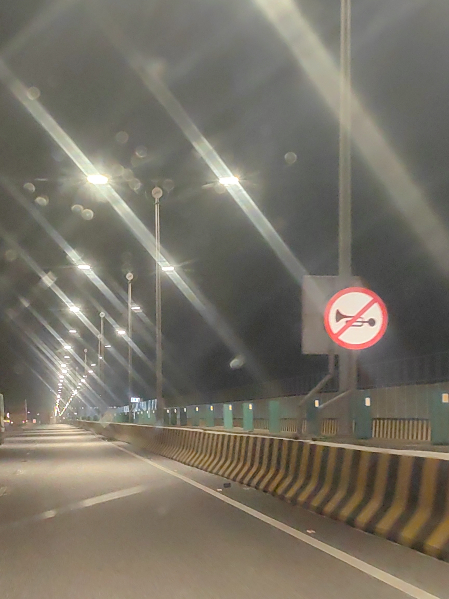 The image captures a nighttime view of a highway in Nhava Khadi, India, characterized by a series of streetlights illuminating the road. A prominent "no honking" sign, a circular red sign with a white background and a black horn symbol crossed out, is mounted on a pole on the right side of the road. The highway features a concrete barrier with alternating yellow and black diagonal stripes, separating the lanes. The road surface is asphalt, with a white lane marking visible. The scene suggests a typical urban or suburban highway environment at night, with artificial lighting and traffic control signage.