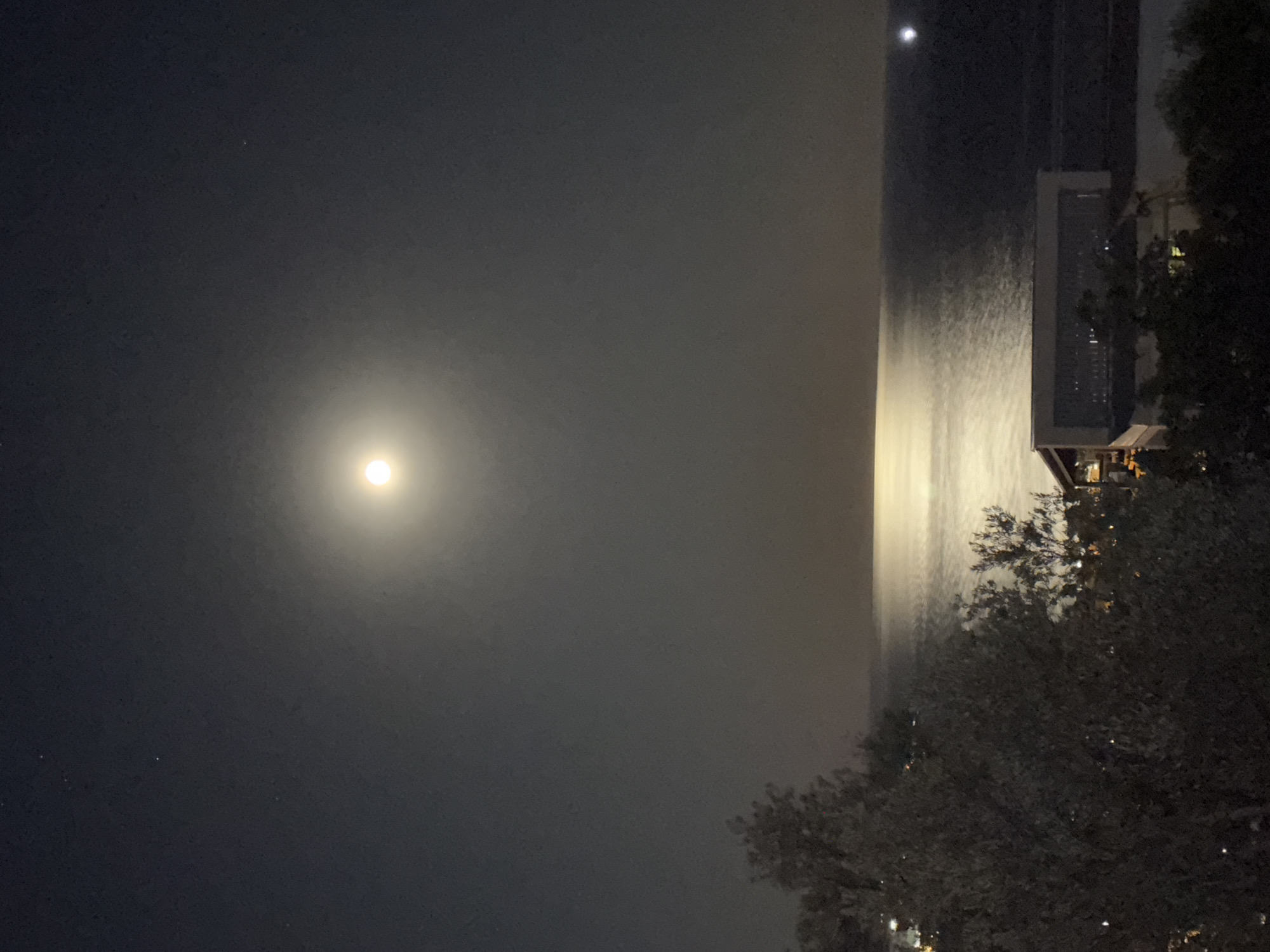 The image shows a night scene in Šušanj, Montenegro, featuring a full moon illuminating the sky and reflecting on the water. A building is visible on the right side, partially obscured by trees. The sky is dark with faint stars, and the water reflects a bright streak of moonlight. The overall environment is calm and quiet.