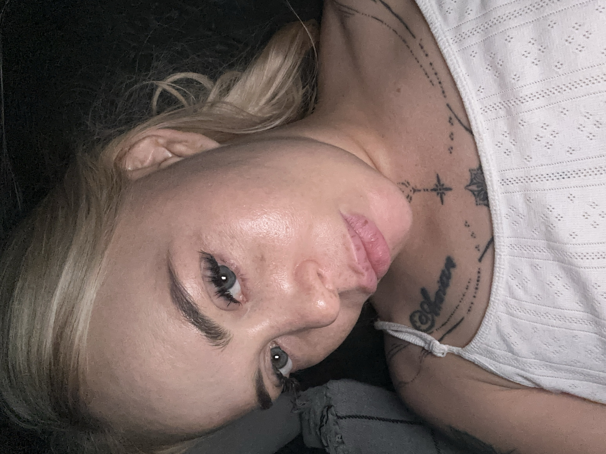 A light-skinned individual, appearing female, is depicted in a close-up, high-angle portrait, looking directly into the camera. Her blonde hair is visible around her head and shoulder. Her eyes are open, with blue irises, and she has prominent dark eyelashes, possibly extended, and dark, well-defined eyebrows. Her skin exhibits visible pores and natural texture, with reflective areas on her forehead, nose, and cheeks, indicative of natural skin oils or illumination. Her lips are full, closed, and have a light pink coloration. She is wearing a white garment, possibly a camisole or tank top, featuring a textured, eyelet or patterned fabric. Multiple tattoos are visible: a script tattoo on her upper left chest/collarbone area, appearing to read "Shania" or similar, accompanied by abstract patterns of lines and dots; centered on her lower neck/upper chest is a small starburst cross design; and further down on her chest, a circular, mandala-like tattoo is partially visible. The background is predominantly dark and out of focus, suggesting low light conditions indoors. A crumpled grey textile object is partially visible on the lower left side of the frame. The overall setting is indoors, in the city of Unknown, Unknown.