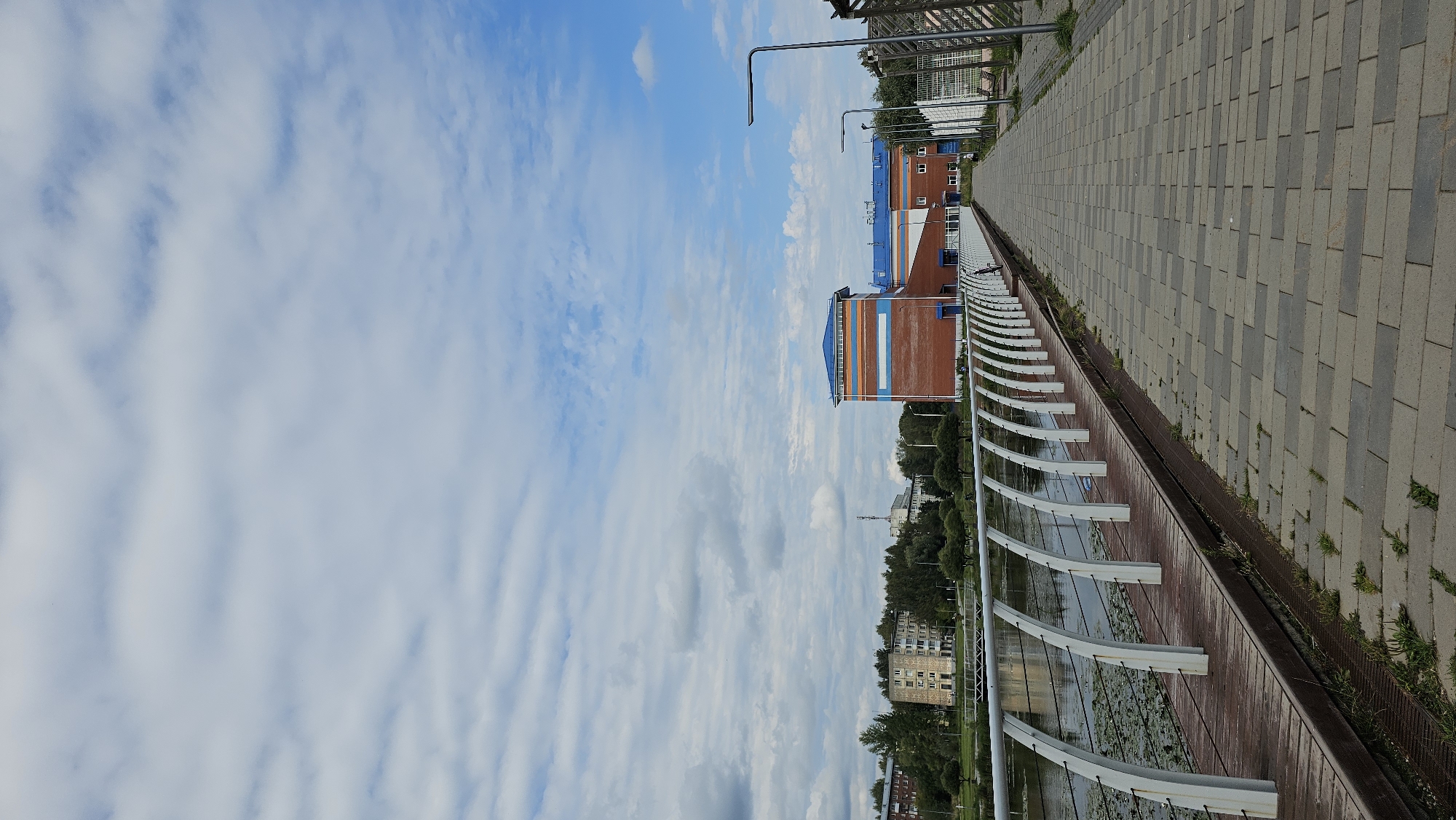 The image depicts a contemporary urban scene in Votkinsk, Russia, likely near a waterfront or park area. In the foreground, a paved walkway extends into the distance, made of grey and beige interlocking rectangular pavers. A modern metal railing with white, curved supports lines the left side of the walkway, separating it from a wooden boardwalk and a body of water. The water, partially covered with lily pads, reflects the cloudy sky and the surrounding buildings.
Beyond the railing and boardwalk, several buildings are visible. A prominent brick building with blue and orange horizontal stripes on its upper facade stands out. To its left, other residential or commercial buildings of varying heights and architectural styles are seen, interspersed with trees. The sky above is a mix of blue sky and white, wispy clouds, indicating fair weather. Streetlights are present along the walkway, suggesting the area is well-maintained for public use. There are no people or animals clearly visible in the frame. The overall impression is one of a developed public space within a city environment.
