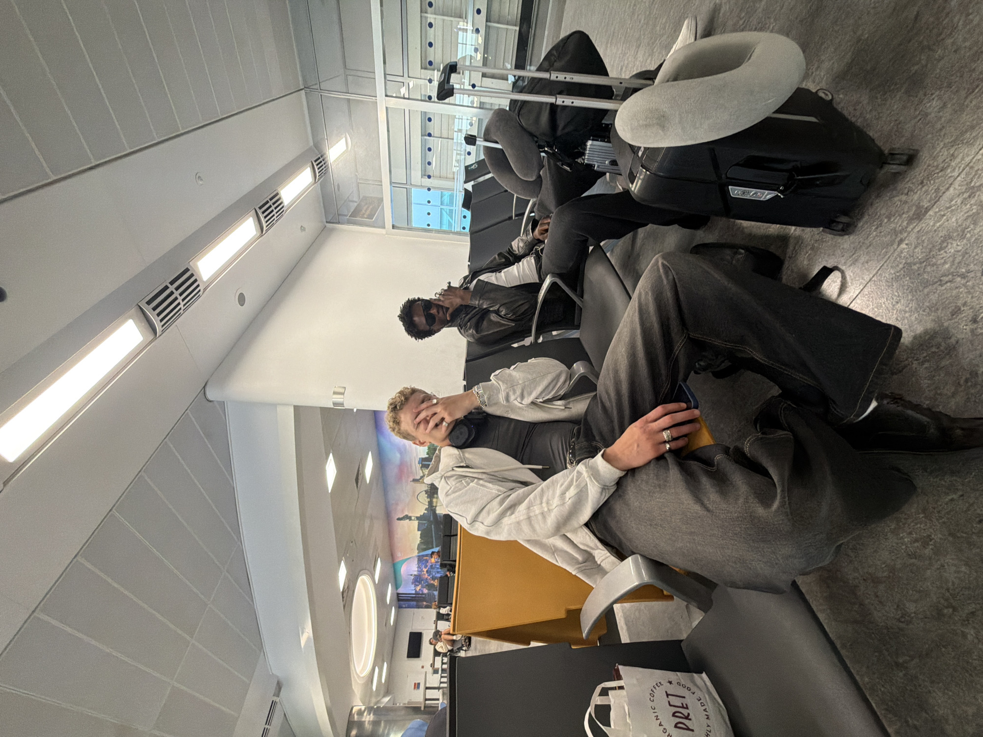 In an airport waiting area, two young men sit in adjacent seats. The man on the left, with blonde hair and a light-colored hoodie, covers his face with his hand and wears headphones around his neck. He has rings on his fingers and is wearing loose-fitting dark denim jeans and a dark top. A white shopping bag with "PRETT" visible on it is on the seat next to him. The man on the right, with dark hair and sunglasses, wears a dark leather jacket over a white tank top and dark pants. He has a cigarette in his hand. Next to him are two suitcases, one of which has a grey neck pillow attached. The airport features a tiled floor, fluorescent lighting, and a mural on the far wall depicting a city skyline. The overall environment suggests a travel terminal.